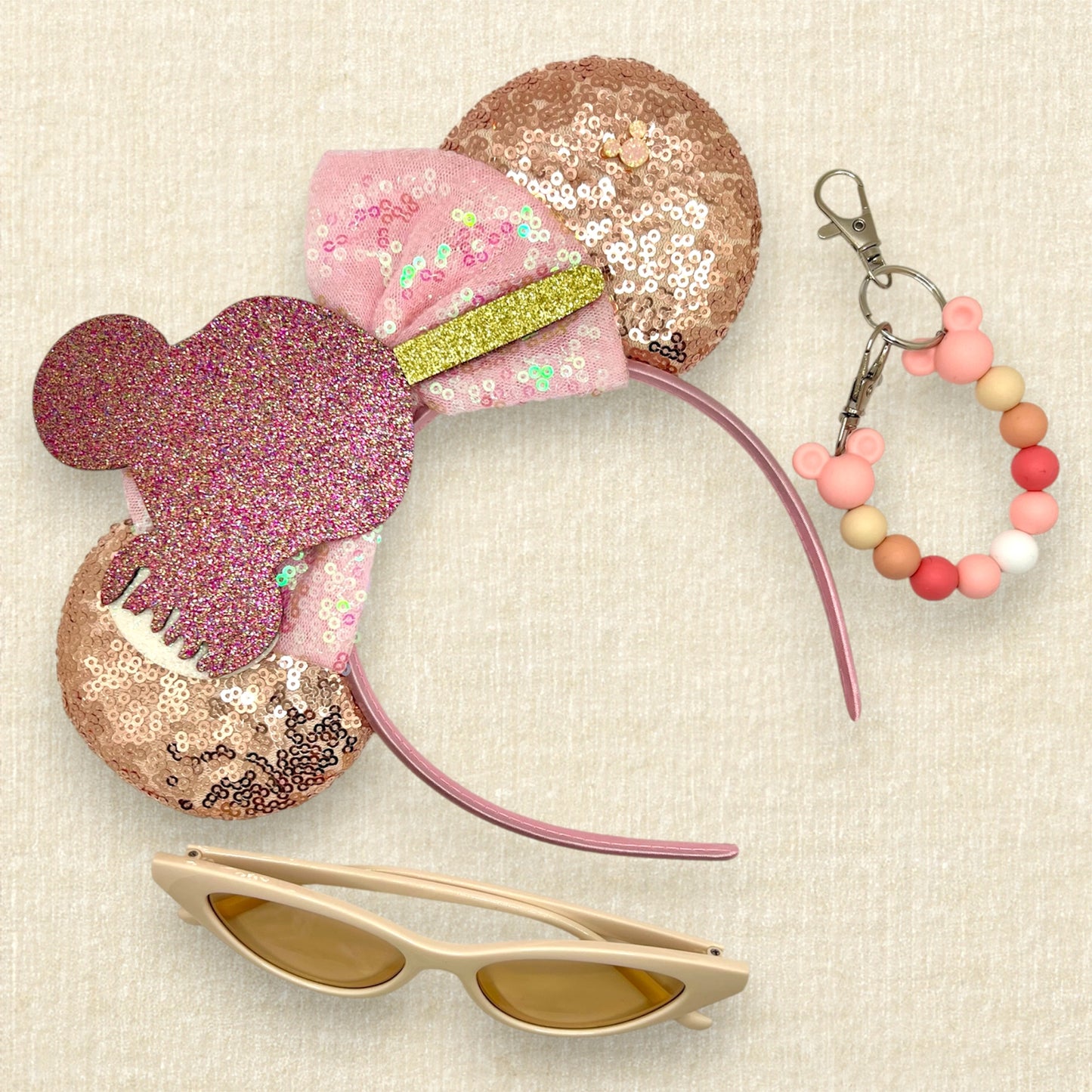 Rose gold sequin mouse ears with iridescent pink bow and glitter mouse-shaped ice cream bar; cream cat-eye sunglasses; pink beaded mouse ears holder keychain
