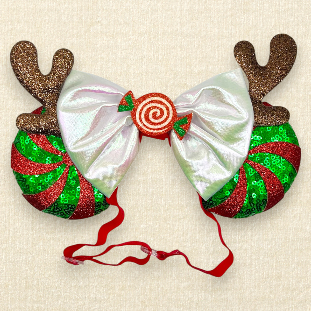 Christmas-themed elastic mouse ears headband with glittery reindeer antlers, red and green peppermint candy cane swirl, an iridescent white bow, and a red spiral candy center on a beige textured background.