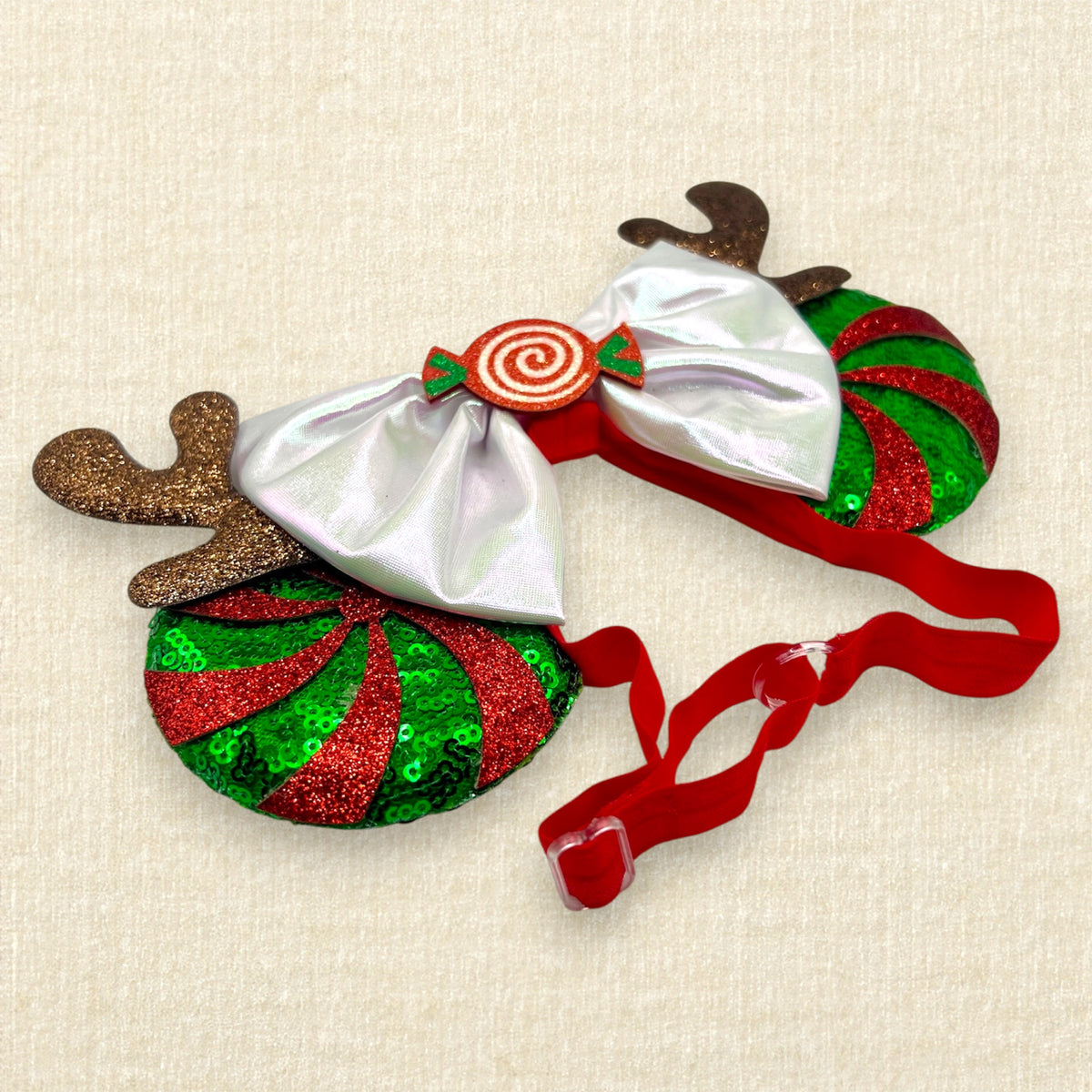 Christmas-themed elastic mouse ears headband with glittery reindeer antlers, red and green peppermint candy cane swirl, an iridescent white bow, and a red spiral candy center on a beige textured background.