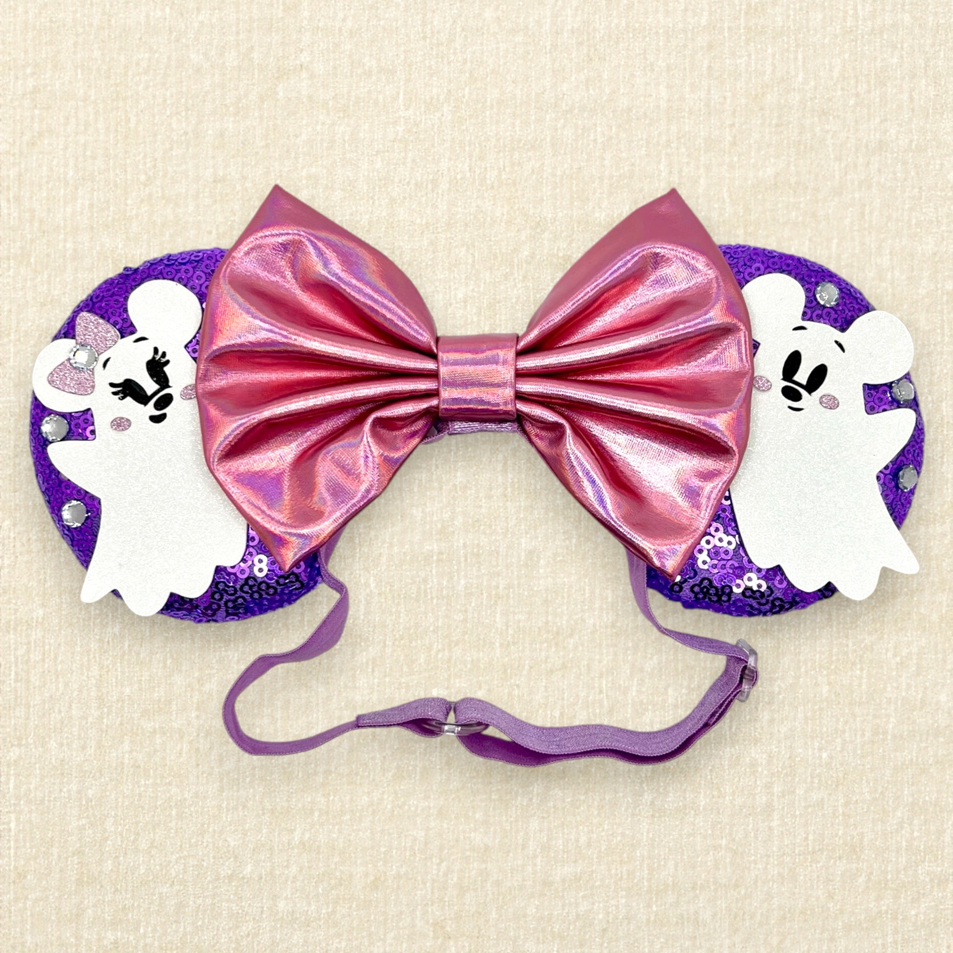 Halloween-themed mouse ears with purple sequin mouse ears, white glitter Mickey & Minnie ghosts, rhinestones, and an iridescent pink bow on an elastic headband