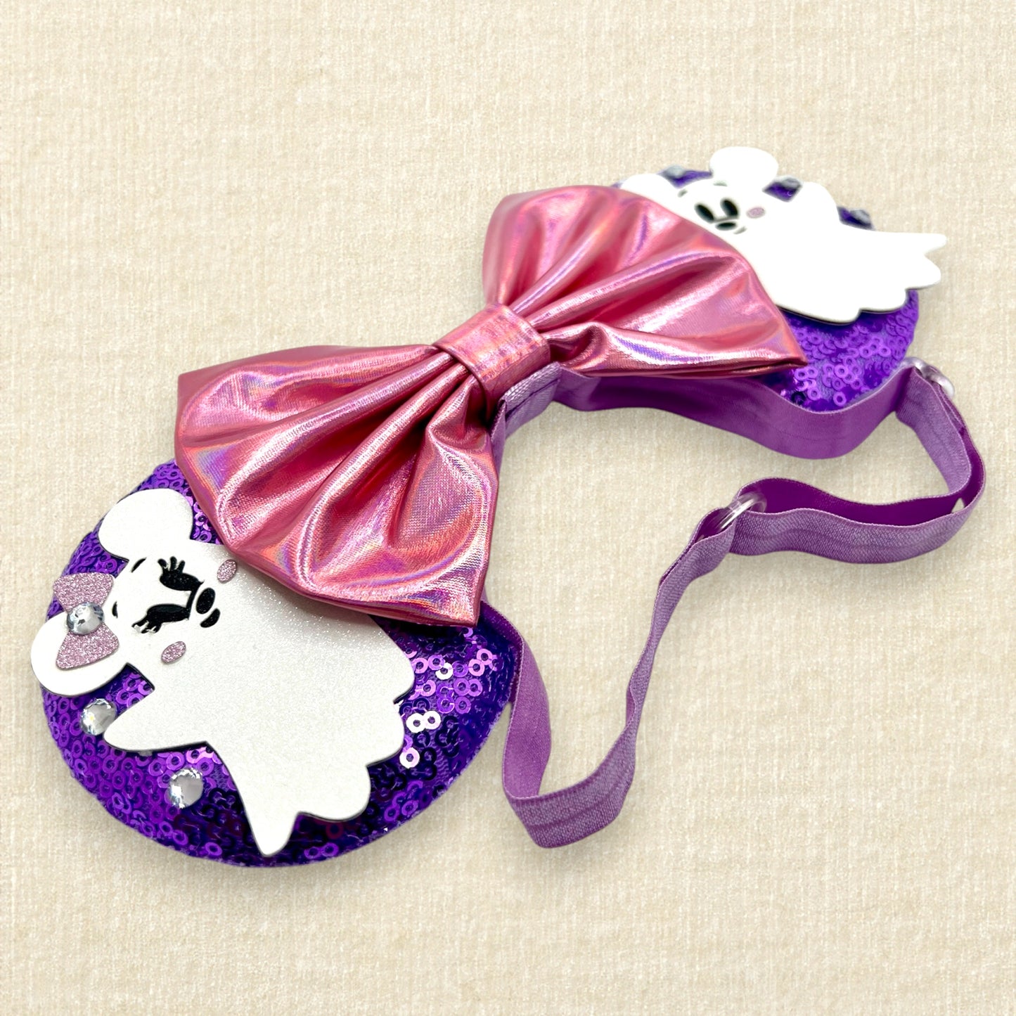 Halloween-themed mouse ears with purple sequin mouse ears, white glitter Mickey & Minnie ghosts, rhinestones, and an iridescent pink bow on an elastic headband