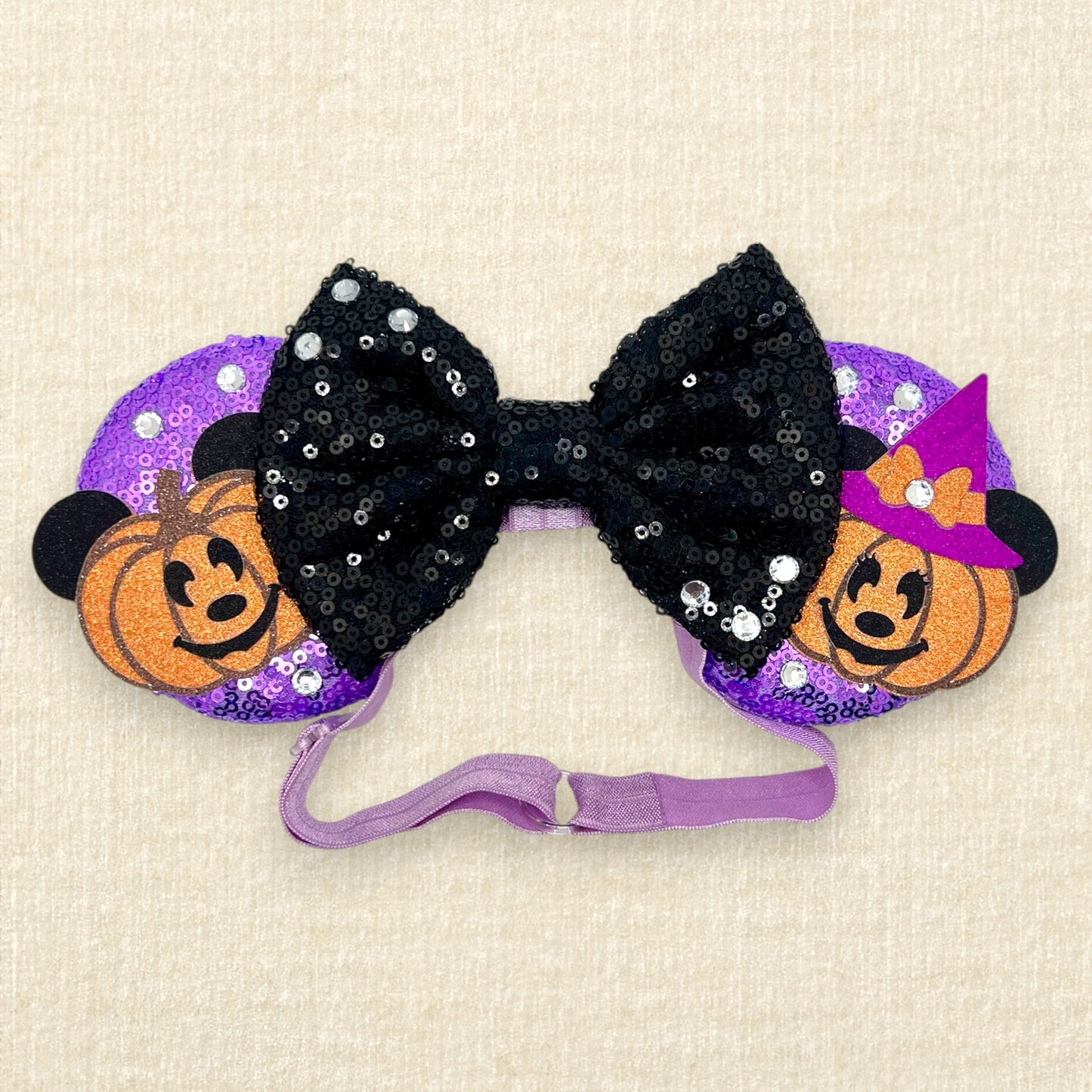 Halloween-themed mouse ears with purple sequin mouse ears, glitter Mickey and Minnie pumpkins, black sequin bow, and rhinestones on an elastic headband