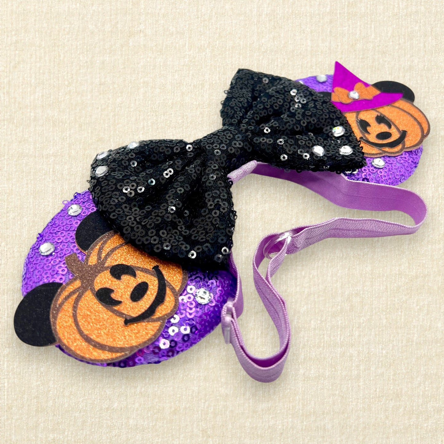 Halloween-themed mouse ears with purple sequin mouse ears, glitter Mickey and Minnie pumpkins, black sequin bow, and rhinestones on an elastic headband