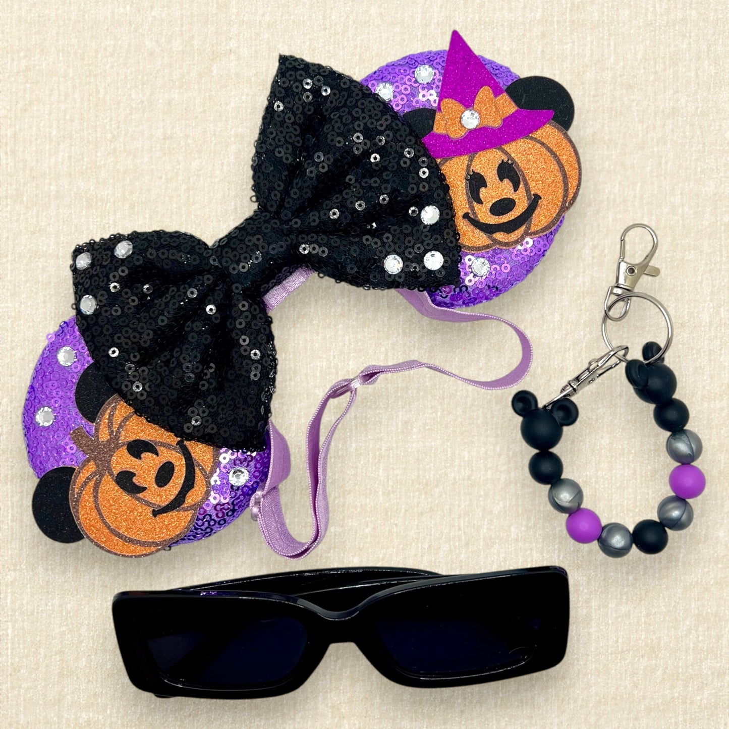 Halloween-themed mouse ears with purple sequin mouse ears, glitter Mickey and Minnie pumpkins, black sequin bow, and rhinestones on an elastic headband; black sunglasses; purple, black, and silver beaded mouse ears holder keychain