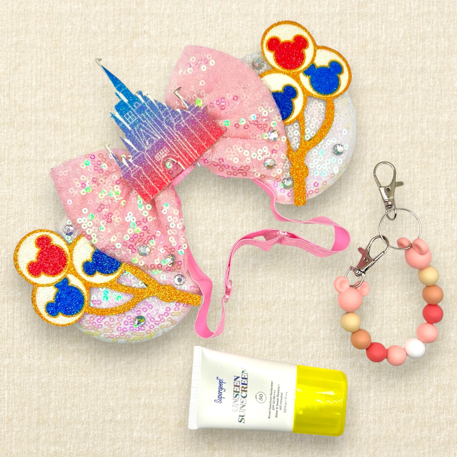 White sequin mouse ears with colorful mouse balloons and a pink bow with pastel pink and blue castle centerpiece on an elastic headband; sunscreen; and pink and white beaded mouse ears holder