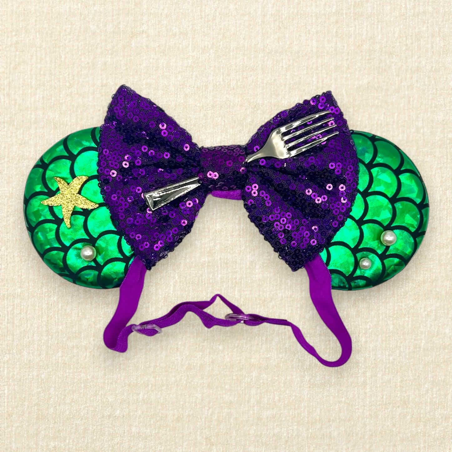 Mouse ears with a green iridescent scale pattern with purple bow, pearls, glitter starfish, and silver fork on an elastic headband