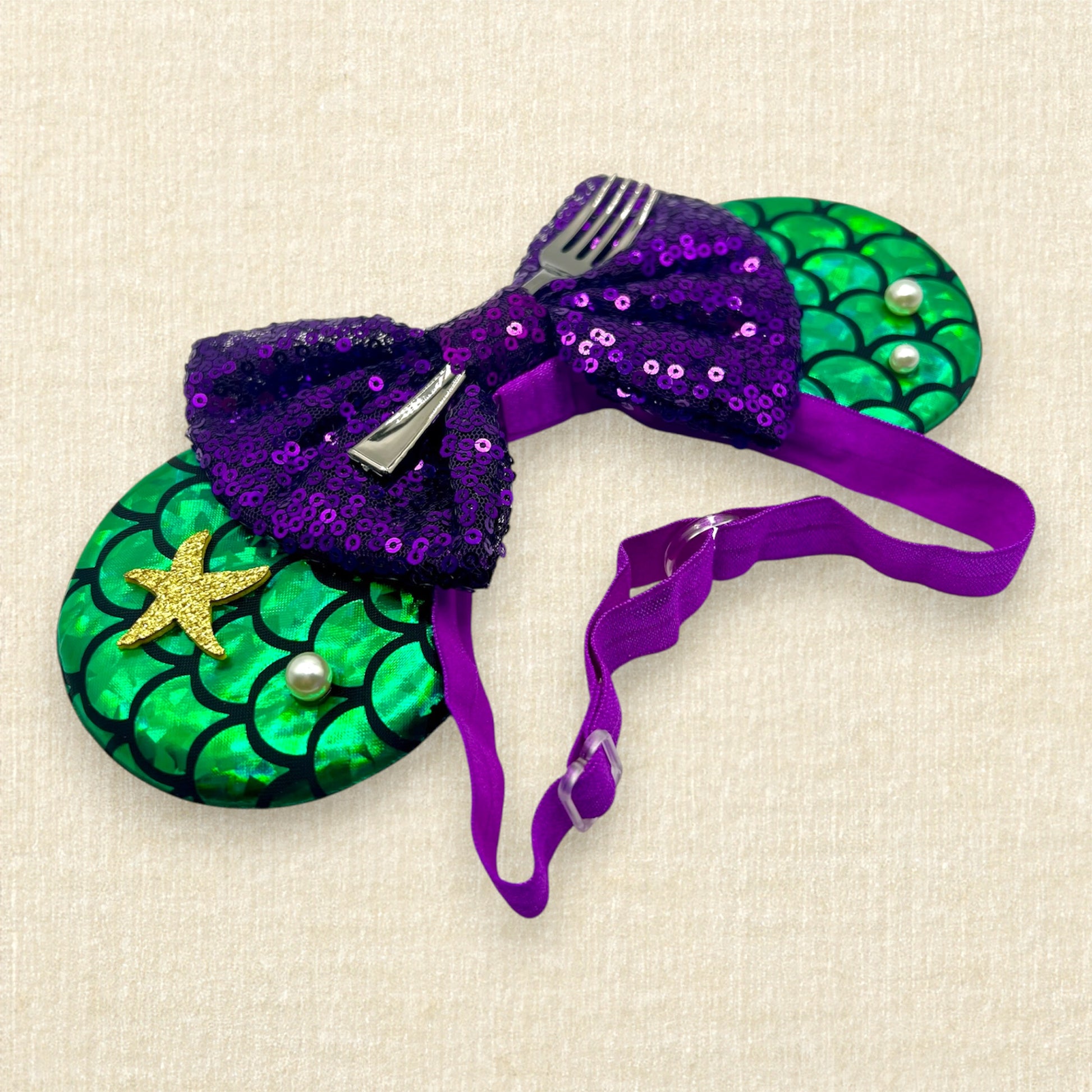 Mouse ears with a green iridescent scale pattern with purple bow, pearls, glitter starfish, and silver fork on an elastic headband