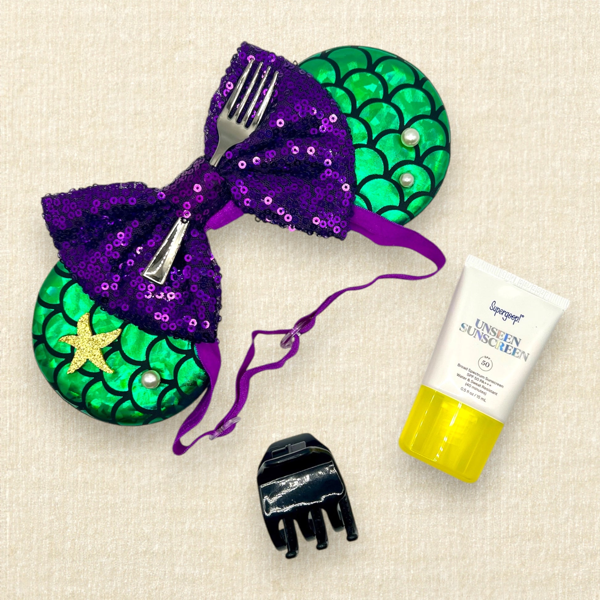 Mouse ears with a green iridescent scale pattern with purple bow, pearls, glitter starfish, and silver fork on an elastic headband; black claw clip; travel sunscreen tube
