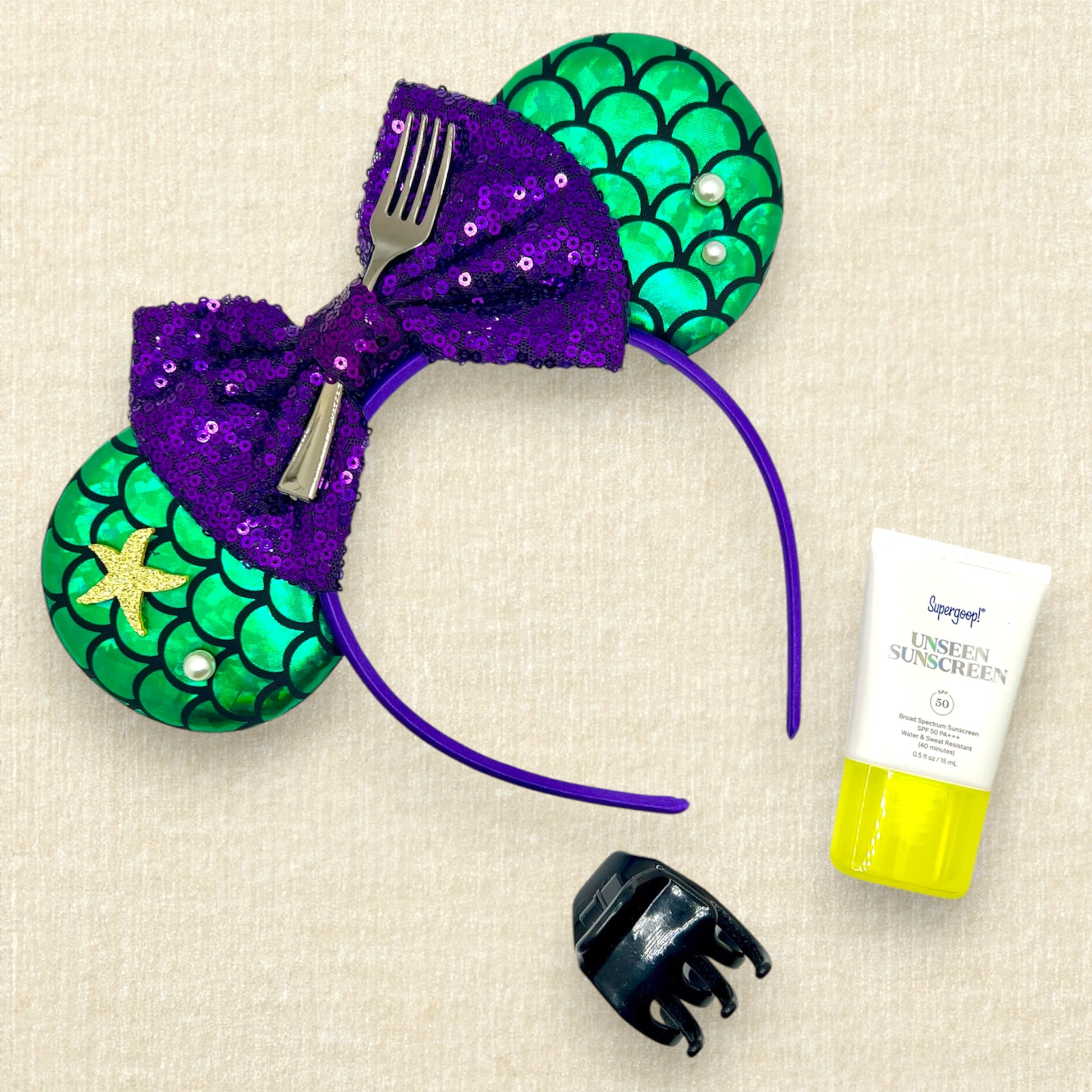 Mouse ears with a green iridescent scale pattern with purple bow, pearls, glitter starfish, and silver fork centerpiece; sunscreen; black claw clip