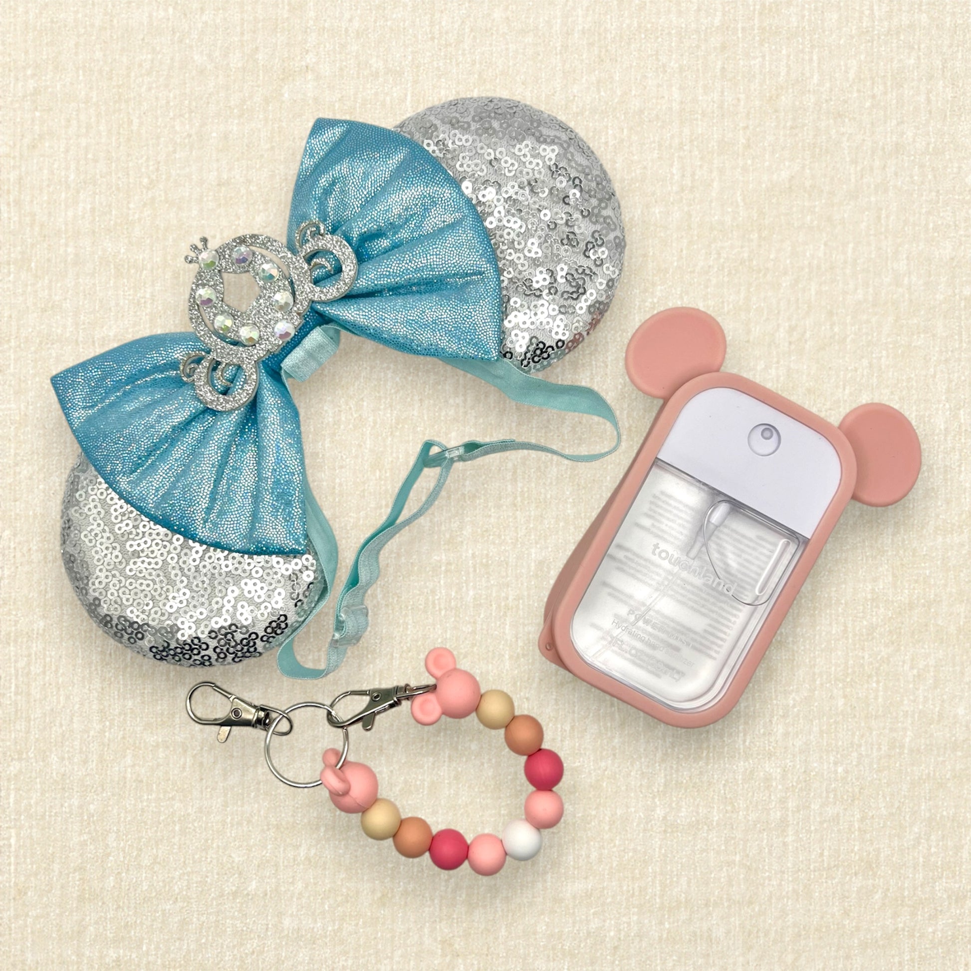 Silver sequin mouse ears on an elastic headband with shimmery blue bow and silver glitter carriage accent and rhinestones; hand sanitizer spray in a pink mouse ears case; pink beaded mouse ears holder clip