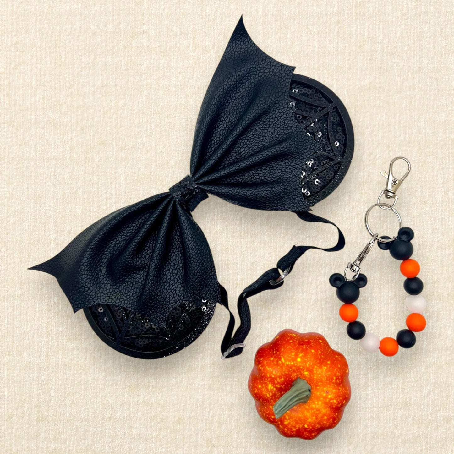 Halloween-themed mouse ears with black sequin ears, black glitter cobwebs, and black faux leather bat wing bow on an elastic headband; small orange pumpkin decoration; orange and black beaded mouse ears holder keychain