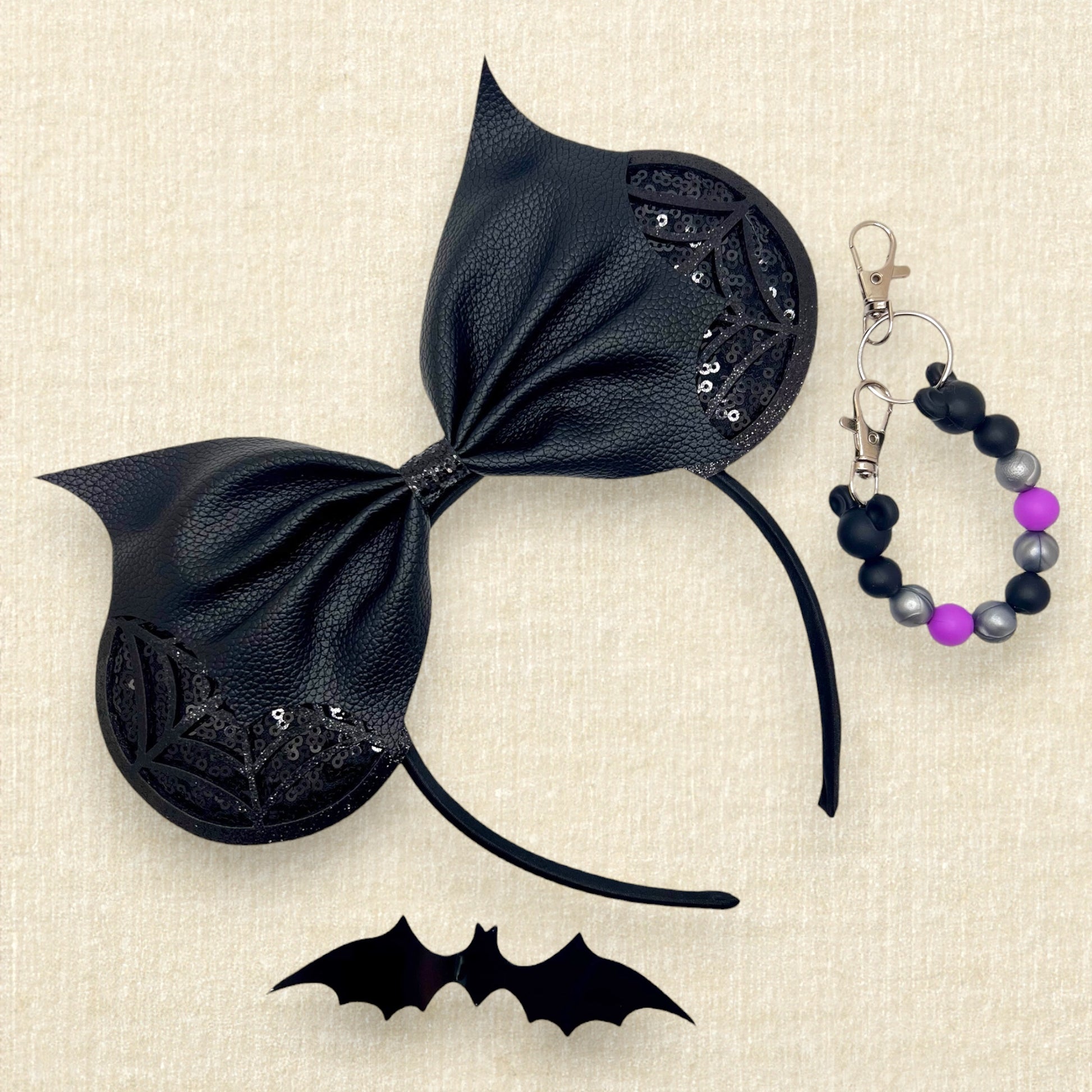 Halloween-themed mouse ears headband with black sequin ears, black glitter cobwebs, and black bat wing faux leather bow; black bat decoration; black, purple, and silver beaded mouse ears holder keychain