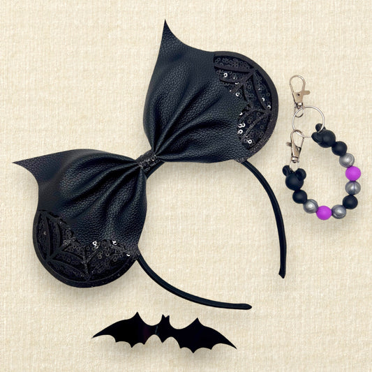 Halloween-themed mouse ears headband with black sequin ears, black glitter cobwebs, and black bat wing faux leather bow; black bat decoration; black, purple, and silver beaded mouse ears holder keychain