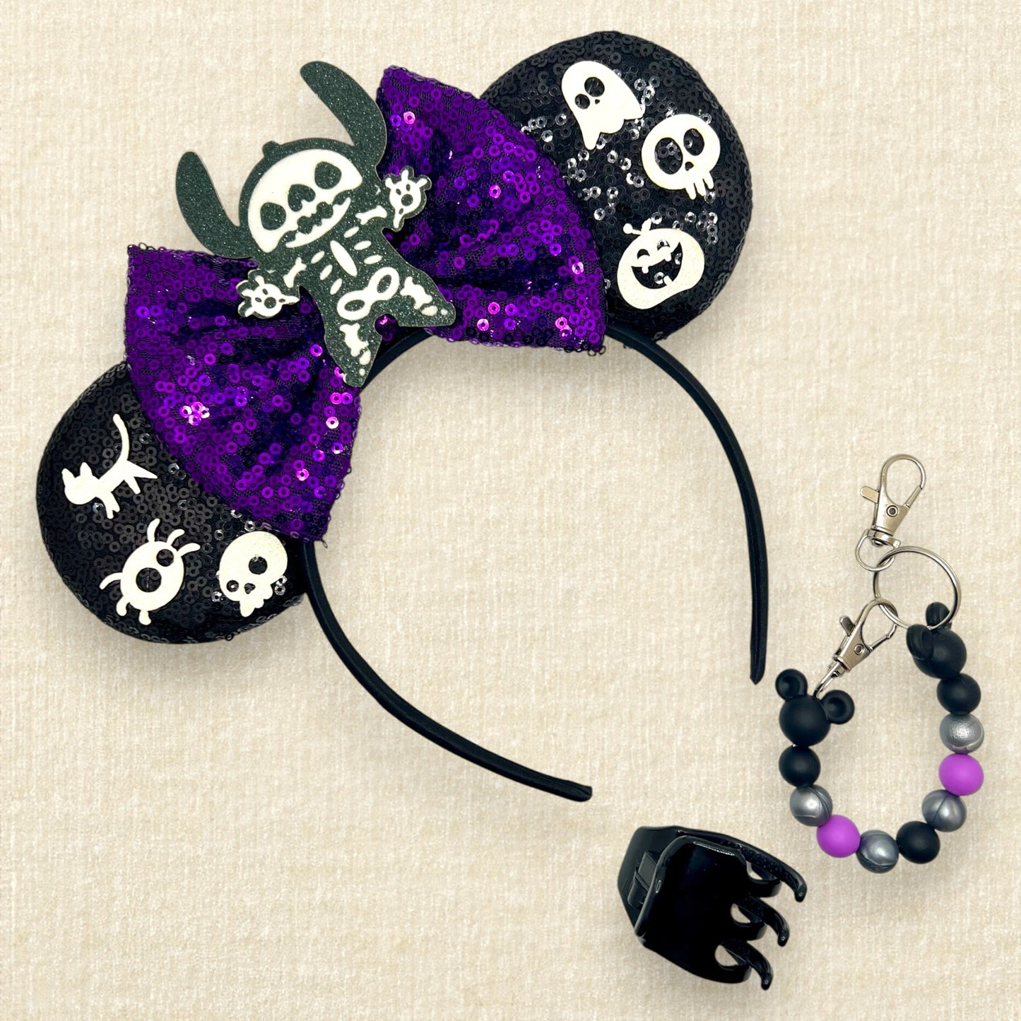 Halloween-themed black mouse ears headband with spooky glitter creatures, purple sequin bow, and skeleton Stitch centerpiece; black claw clip; purple, black, and silver beaded mouse ears holder keychain