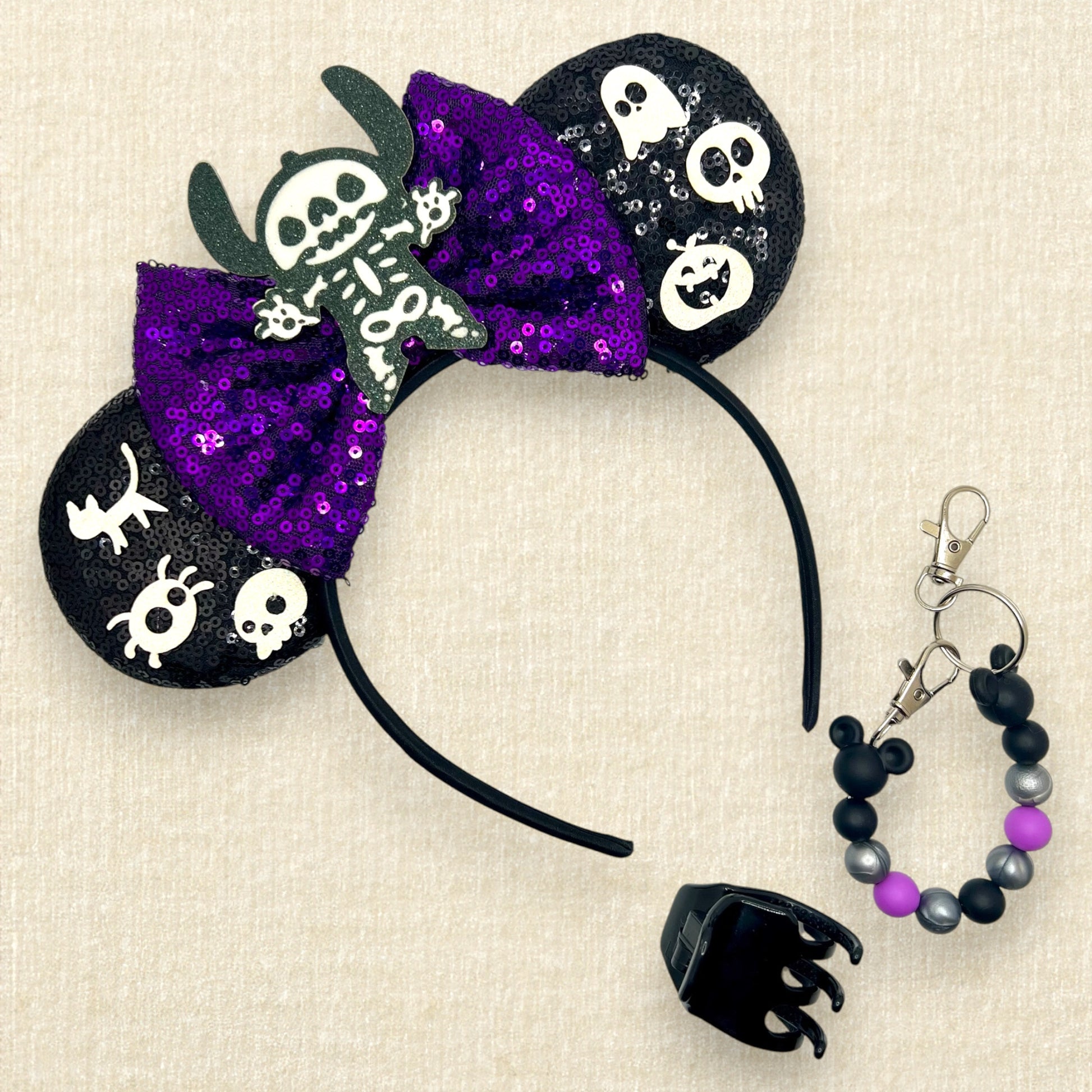 Halloween-themed black mouse ears headband with spooky glitter creatures, purple sequin bow, and skeleton Stitch centerpiece; black claw clip; purple, black, and silver beaded mouse ears holder keychain