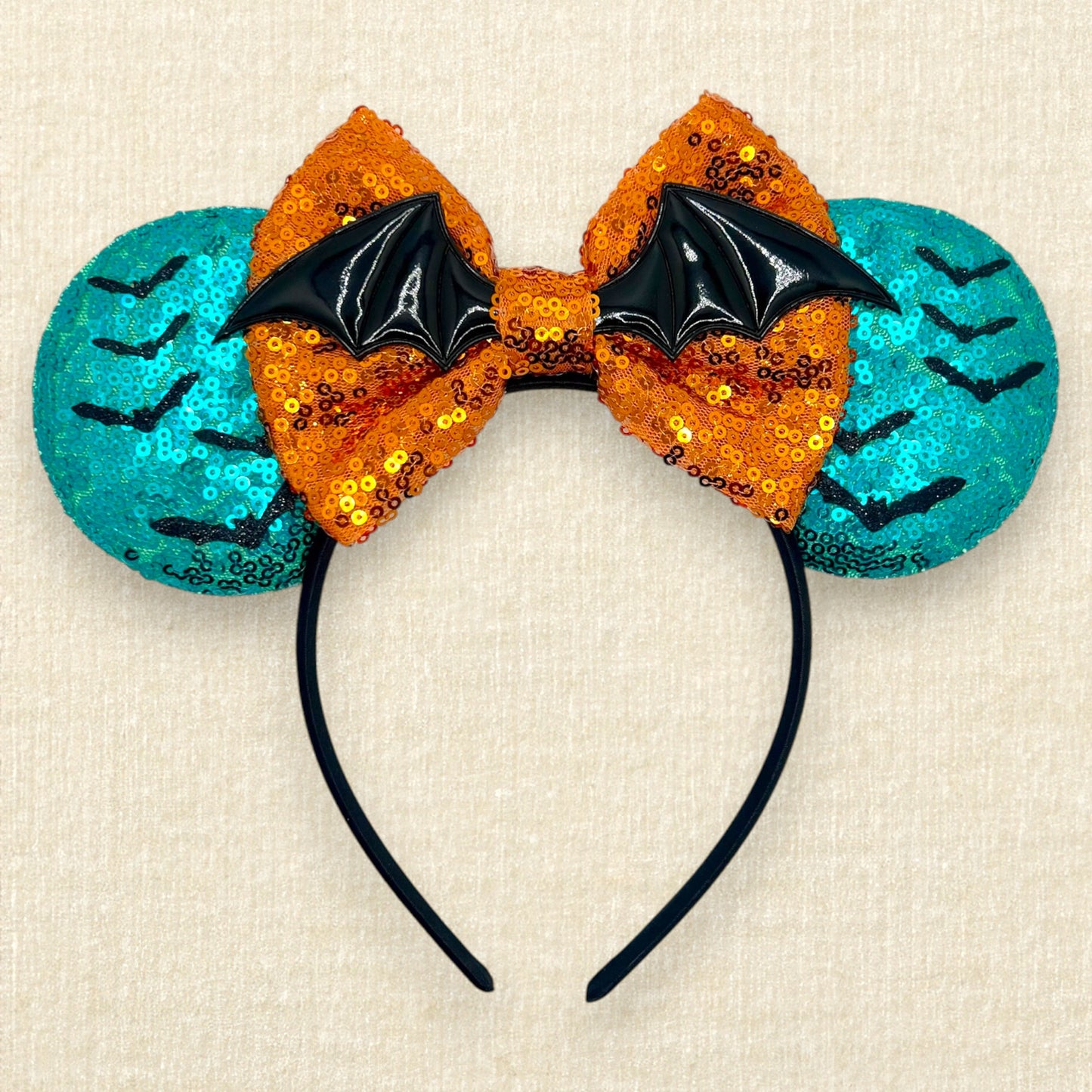 Halloween-themed mouse ears headband with orange sequin bow, black bats, and teal blue ears on a beige background