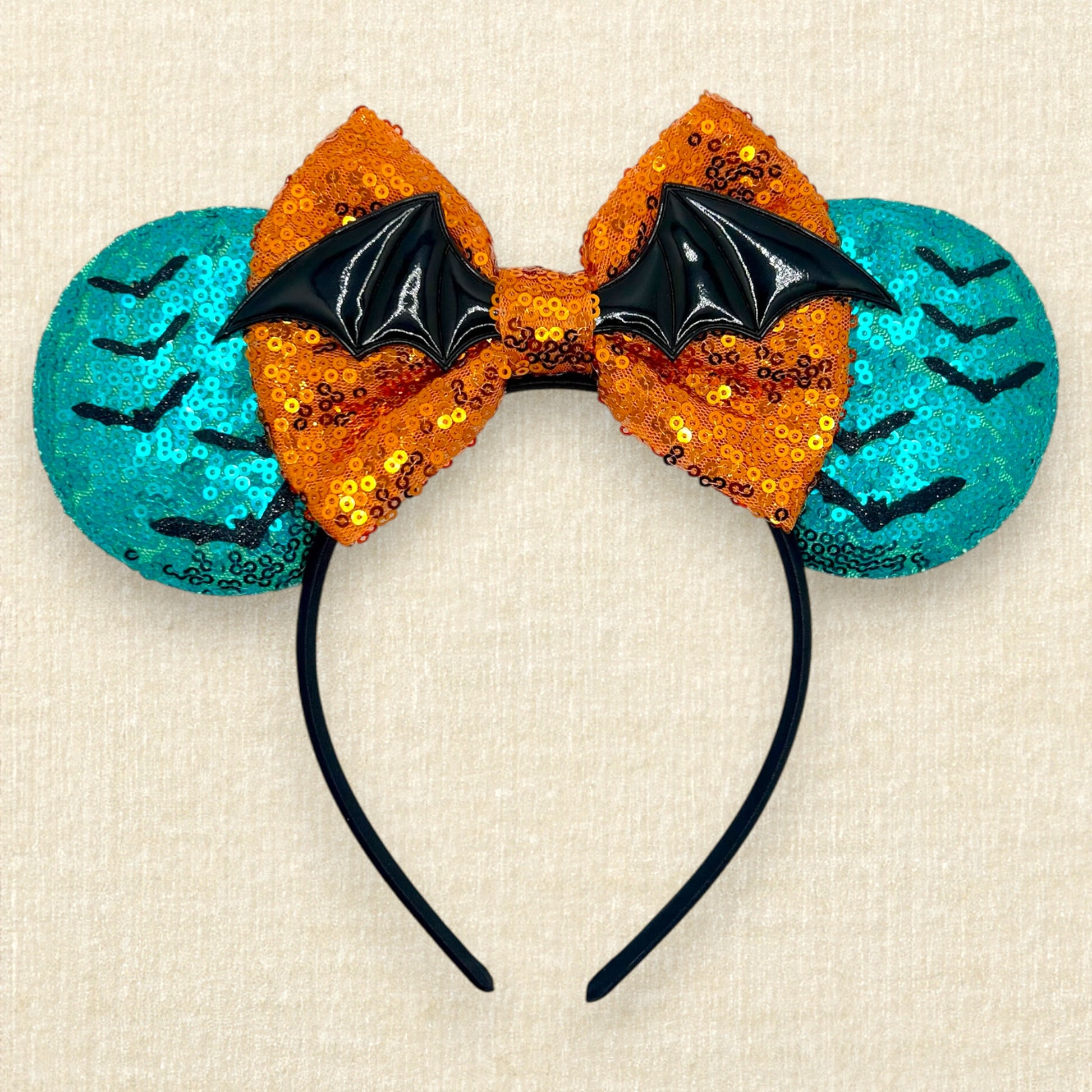 Halloween-themed mouse ears headband with orange sequin bow, black bats, and teal blue ears on a beige background