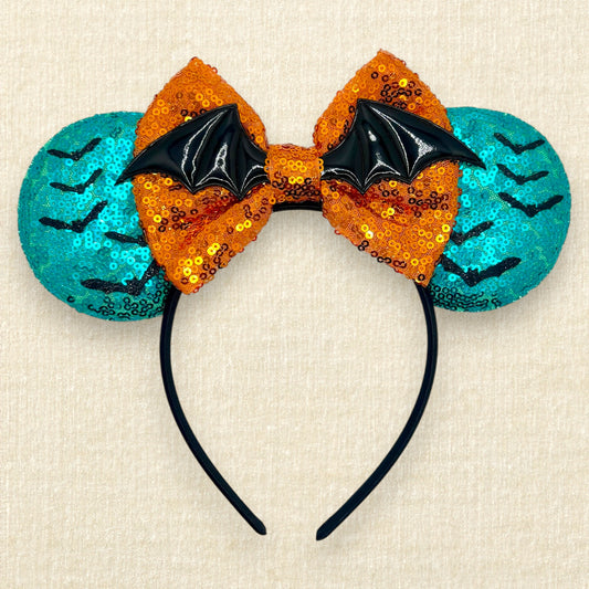 Halloween-themed mouse ears headband with orange sequin bow, black bats, and teal blue ears on a beige background