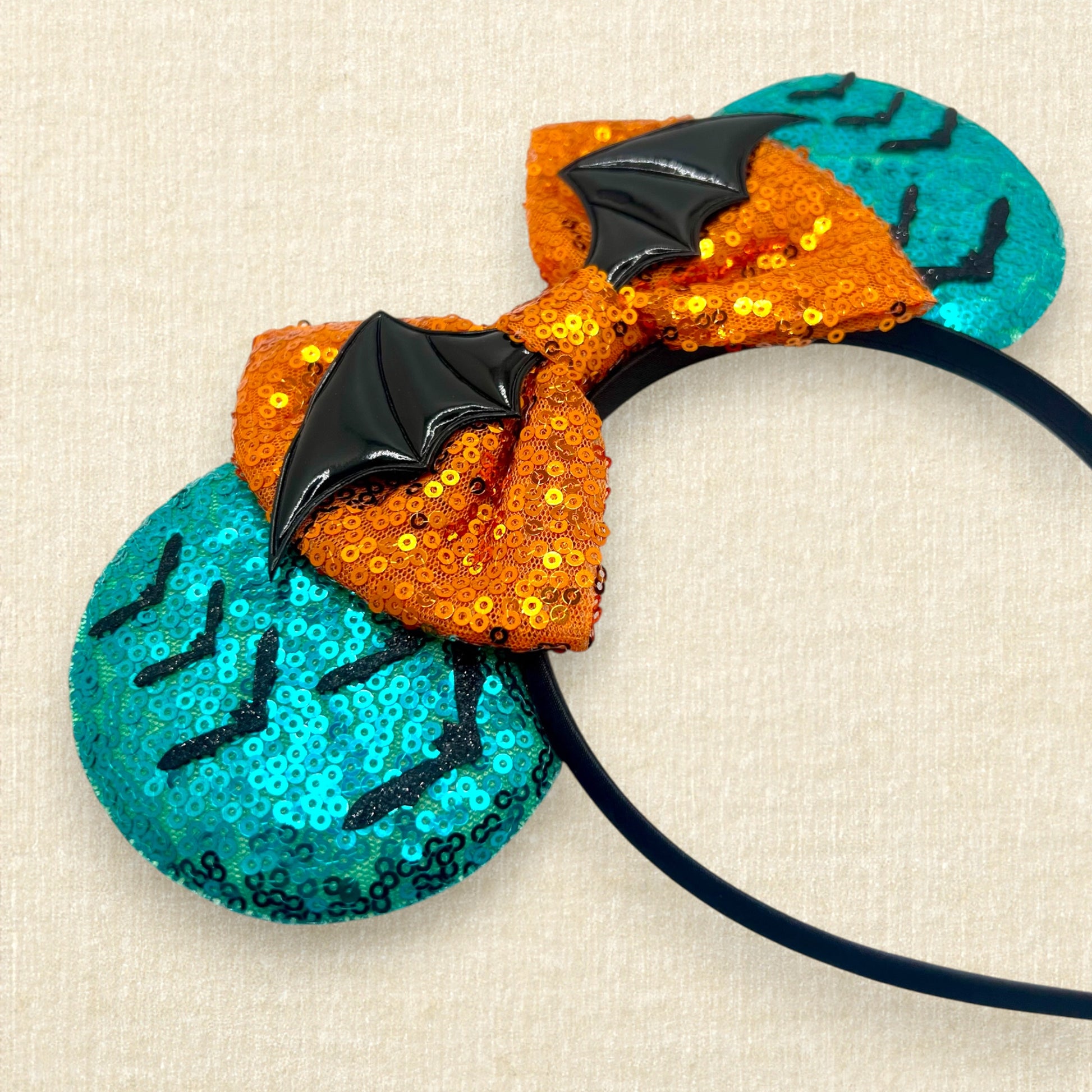 Halloween-themed mouse ears headband with orange sequin bow, black bats, and teal blue ears on a beige background
