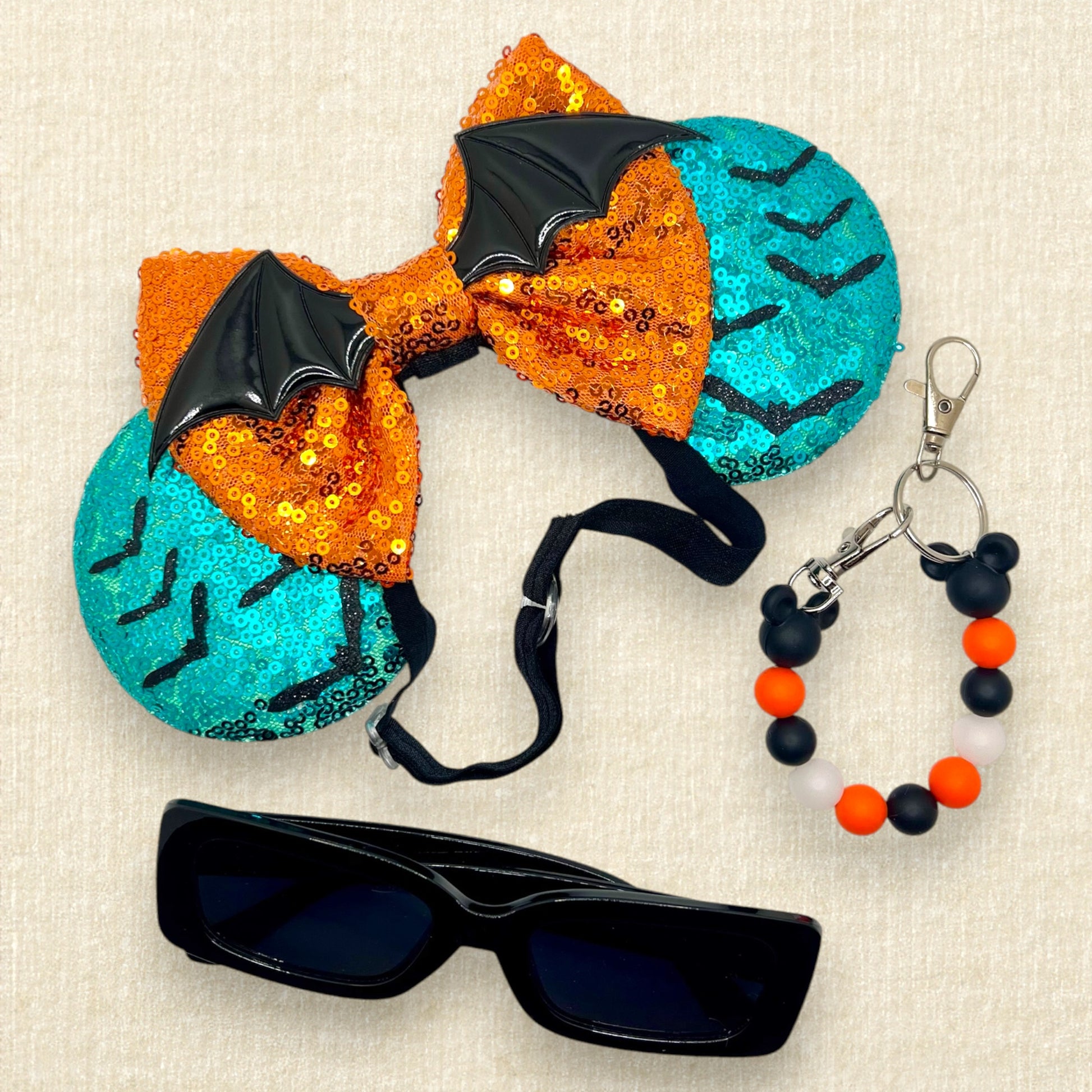 Halloween-themed teal mouse ears with black bats, orange bow, and large bat wings; black sunglasses; black and orange beaded mouse ears holder keychain
