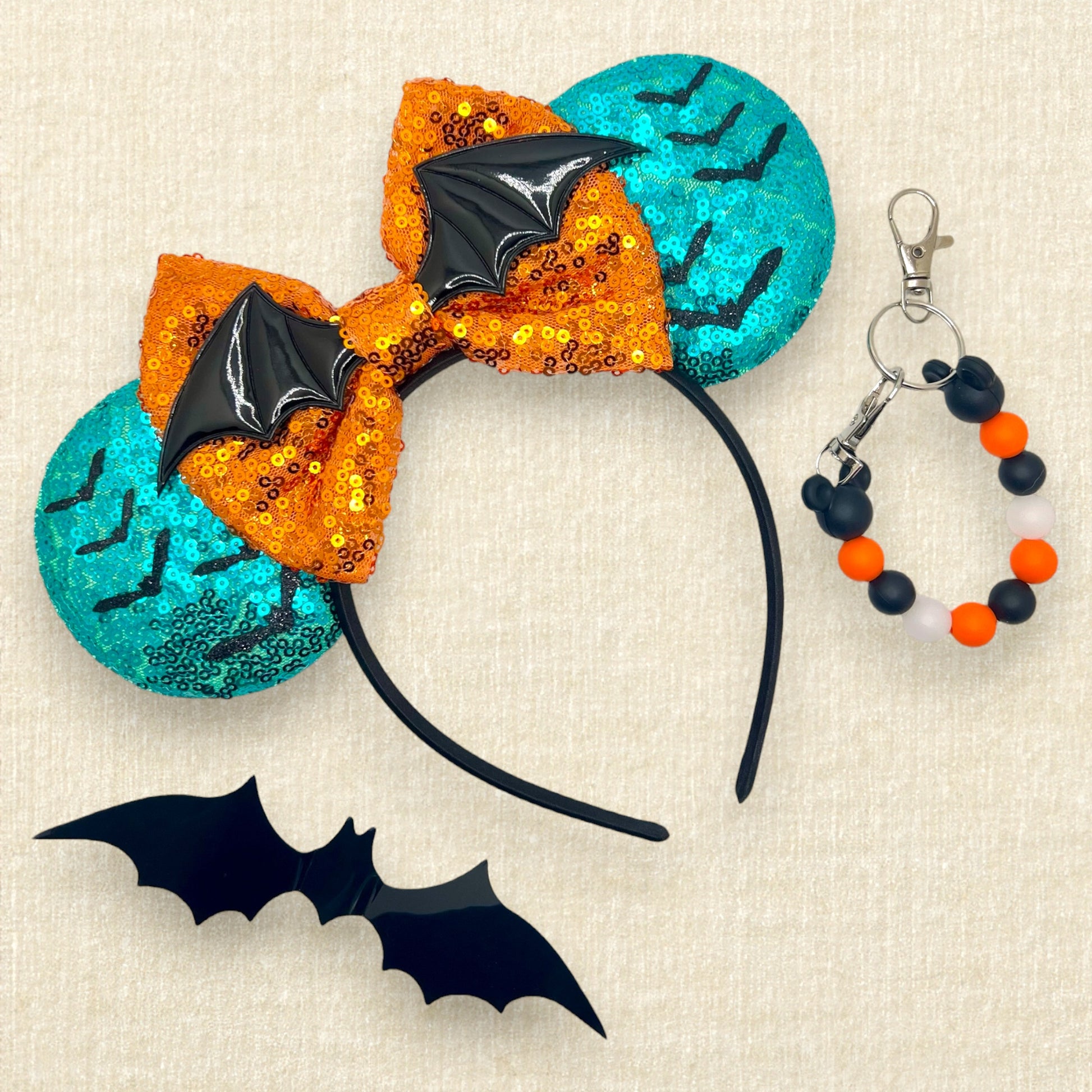 Halloween-themed mouse ears headband with orange sequin bow, black bats, and teal blue ears on a beige background; black bat decoration; orange and black beaded mouse ears holder keychain