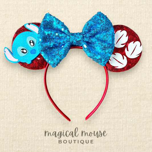 Mouse ear headband with blue bow and red mouse ears with a mini blue alien and white palm leaves on a beige background, branded 'magical mouse boutique'.