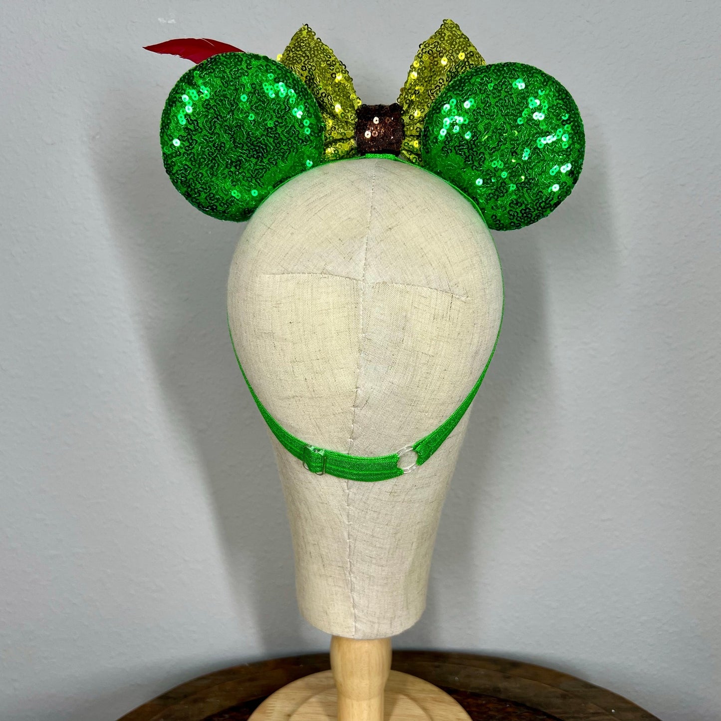 Green sequin elastic mouse ears  with lighter green sequin bow and red feather headband on a mannequin with a gray background