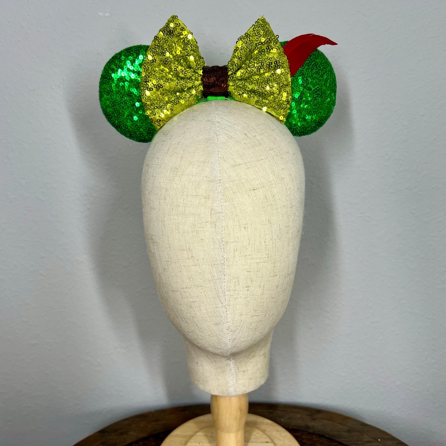 Green sequin elastic mouse ears  with lighter green sequin bow and red feather headband on a mannequin with a gray background