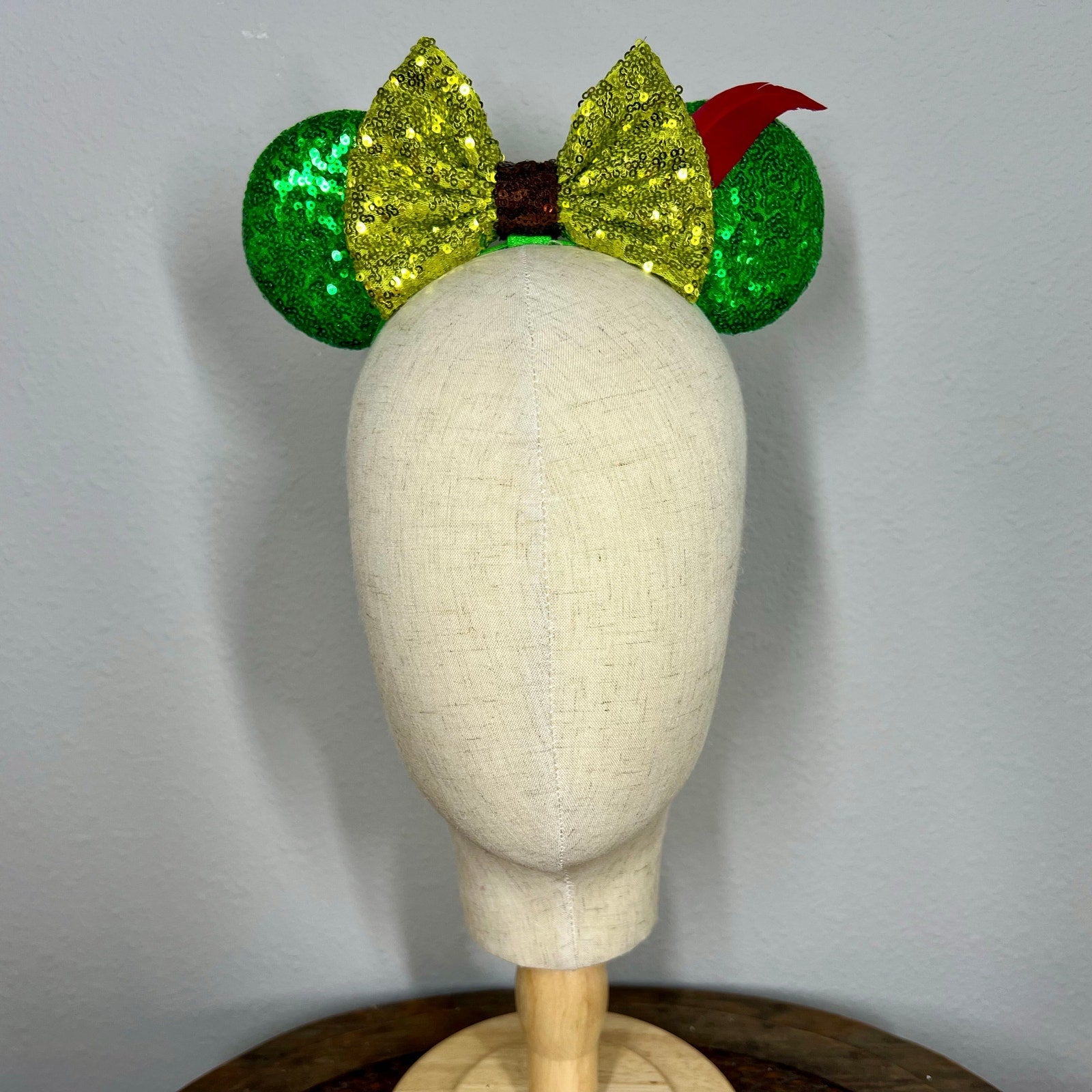 Green sequin elastic mouse ears  with lighter green sequin bow and red feather headband on a mannequin with a gray background
