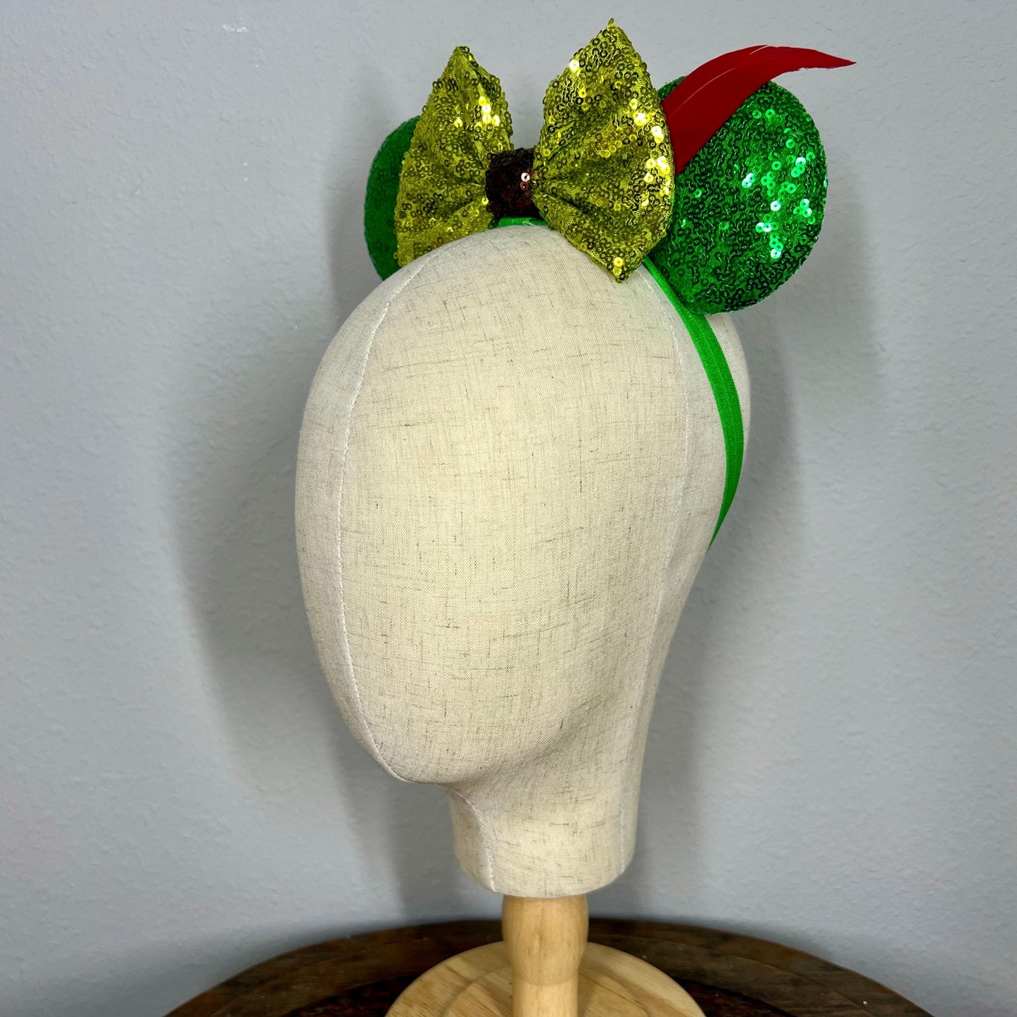 Green sequin elastic mouse ears  with lighter green sequin bow and red feather headband on a mannequin with a gray background
