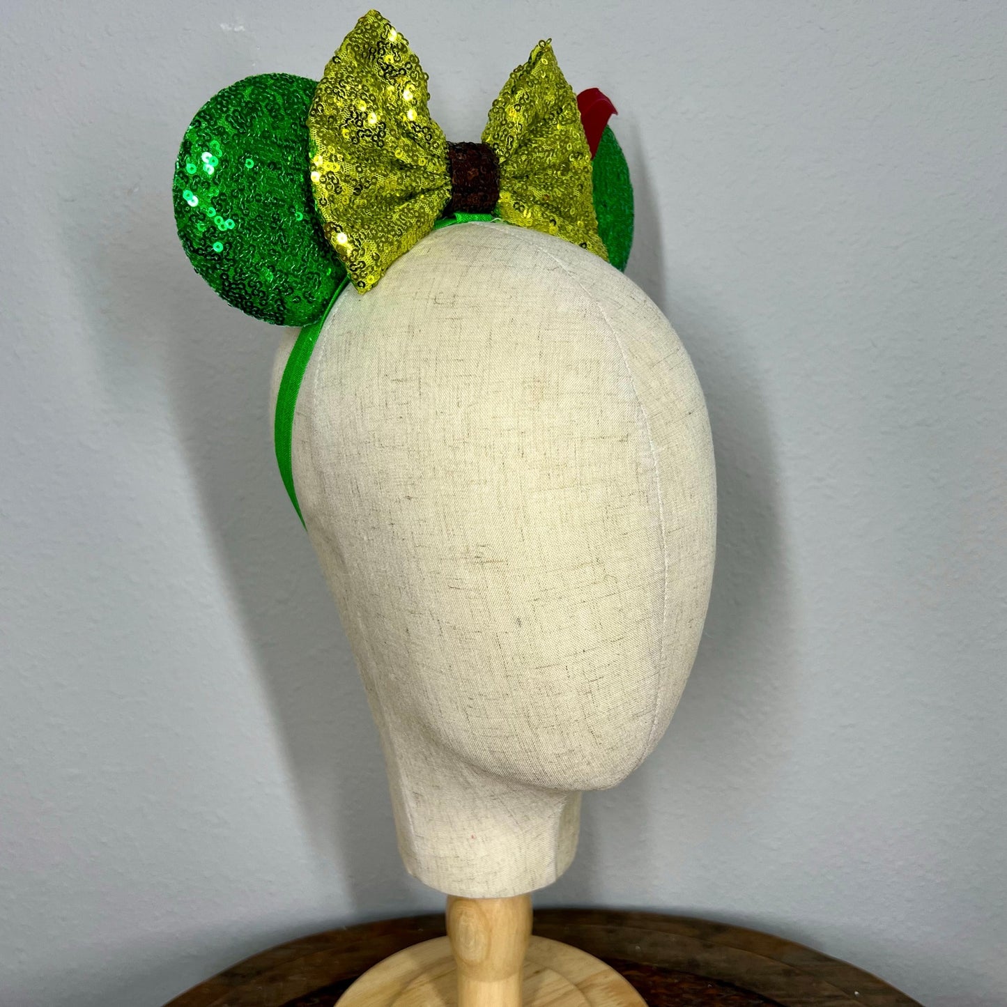 Green sequin elastic mouse ears  with lighter green sequin bow and red feather headband on a mannequin with a gray background