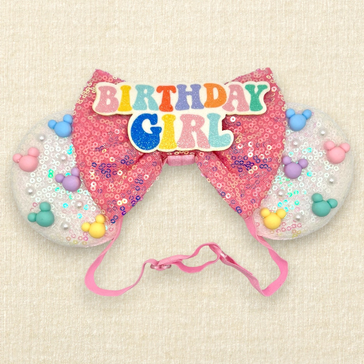 Iridescent white mouse ears with colorful mouse-shaped pearls, pink sequin bow, and glitter 'Birthday Girl' text centerpiece on an elastic headband