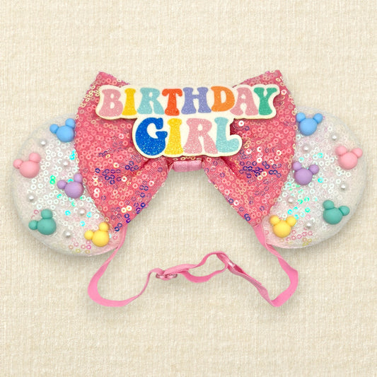 Iridescent white mouse ears with colorful mouse-shaped pearls, pink sequin bow, and glitter 'Birthday Girl' text centerpiece on an elastic headband