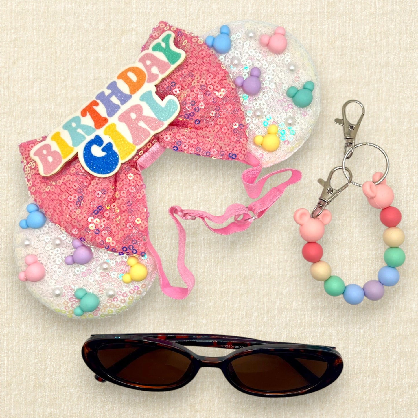 Iridescent white mouse ears with colorful mouse-shaped pearls, pink sequin bow, and glitter 'Birthday Girl' text centerpiece on an elastic headband; tortoiseshell sunglasses; coloful beaded mouse ears holder keychain