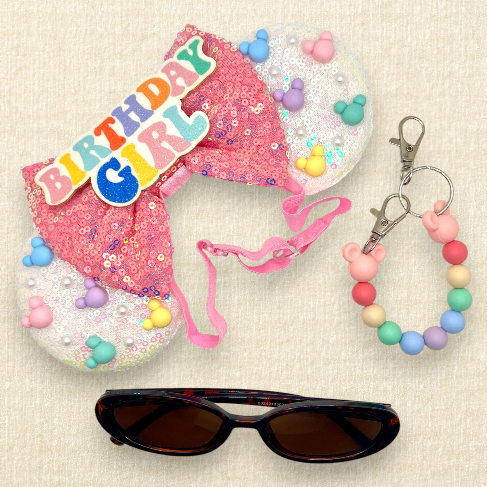 Iridescent white mouse ears with colorful mouse-shaped pearls, pink sequin bow, and glitter 'Birthday Girl' text centerpiece on an elastic headband; tortoiseshell sunglasses; coloful beaded mouse ears holder keychain