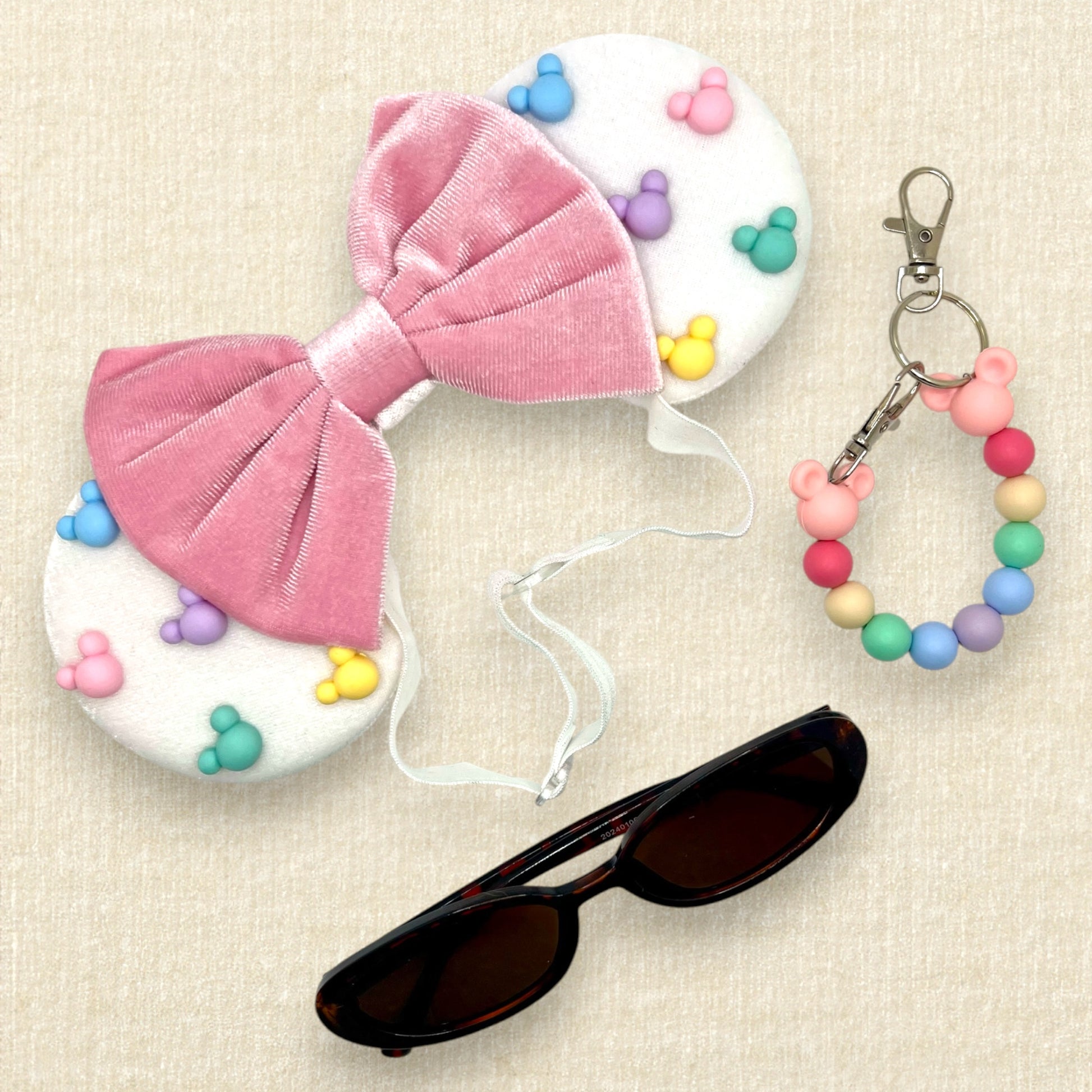White velvet mouse ears with colorful pastel mouse pearls and pastel pink velvet bow on an elastic headband; tortoiseshell sunglasses; colorful rainbow beaded mouse ears holder keychain