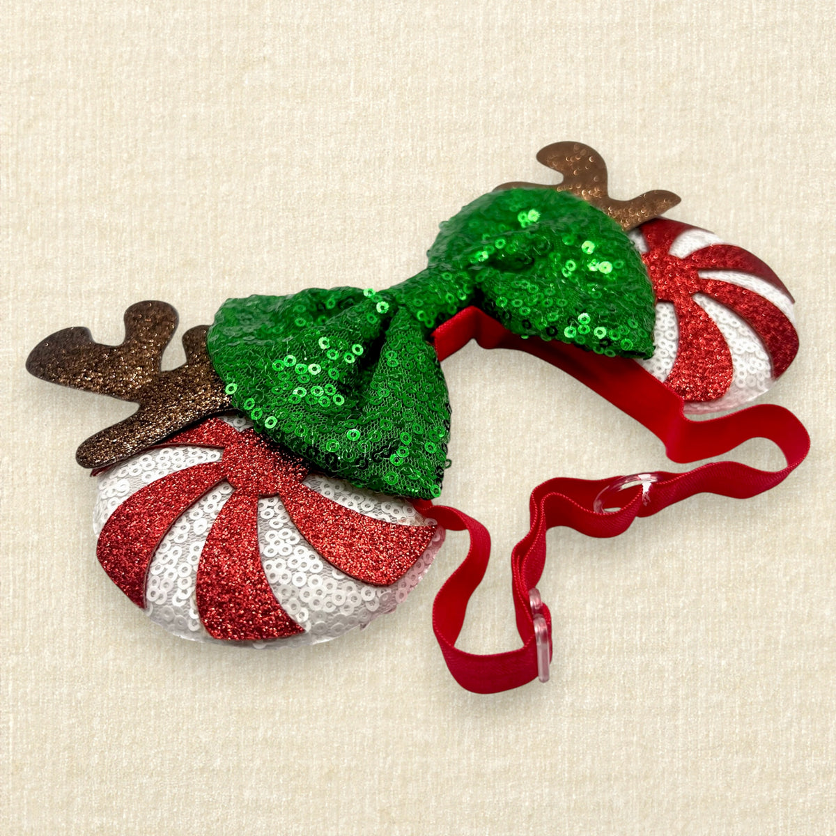 Christmas-themed mouse ears headband with a large green sequined bow, brown reindeer antlers, and candy cane side mouse ears in red and white stripes, shown on a light beige textured background.
