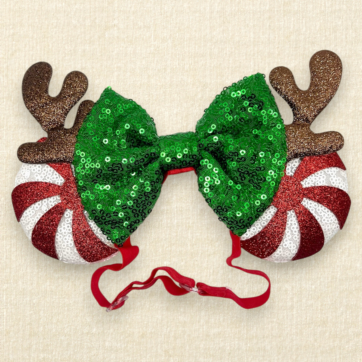 Christmas-themed mouse ears headband with a large green sequined bow, brown reindeer antlers, and candy cane side mouse ears in red and white stripes, shown on a light beige textured background.