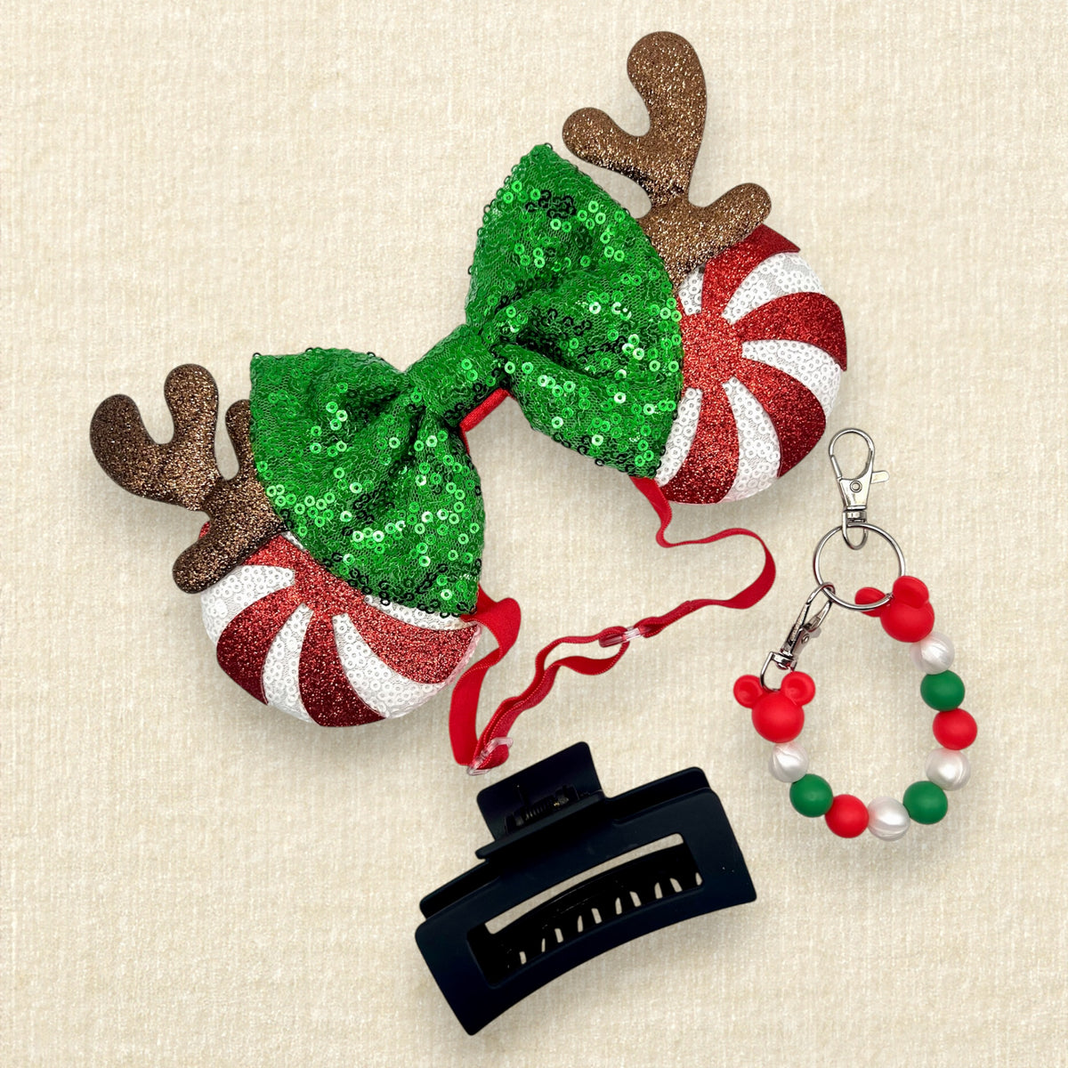 Christmas-themed mouse ears headband with a large green sequined bow, brown reindeer antlers, and candy cane side mouse ears in red and white stripes; a red, green, and pearl white beaded mouse ears holder keychain clip; and a black claw clip.