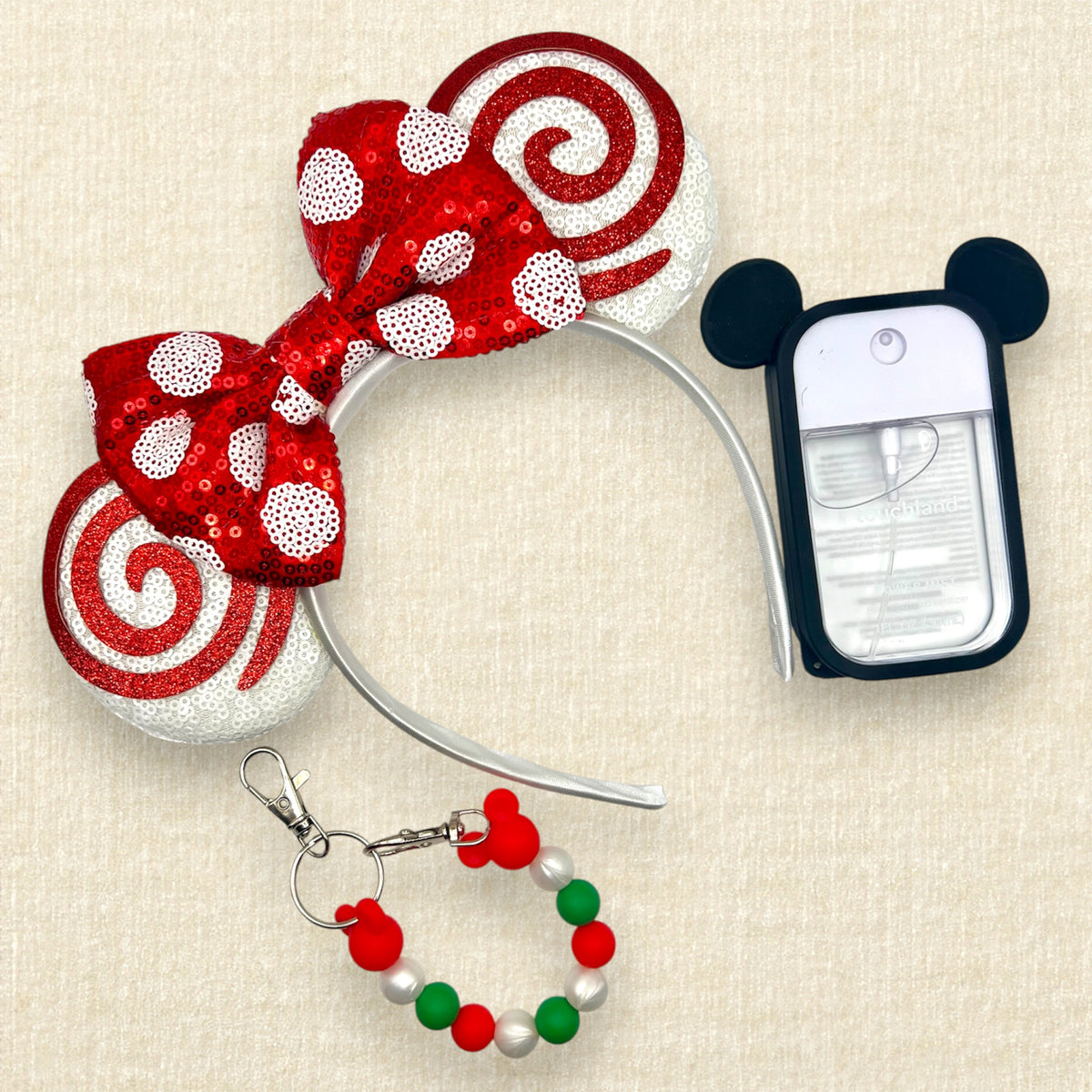 A festive Minnie Mouse-inspired headband accessory set on a textured beige background, featuring a red and white sequined bow and ears, a small red, green, and white beaded mouse ears holder keychain clip, and a black-and-white hand sanitizer spray with mouse ears.