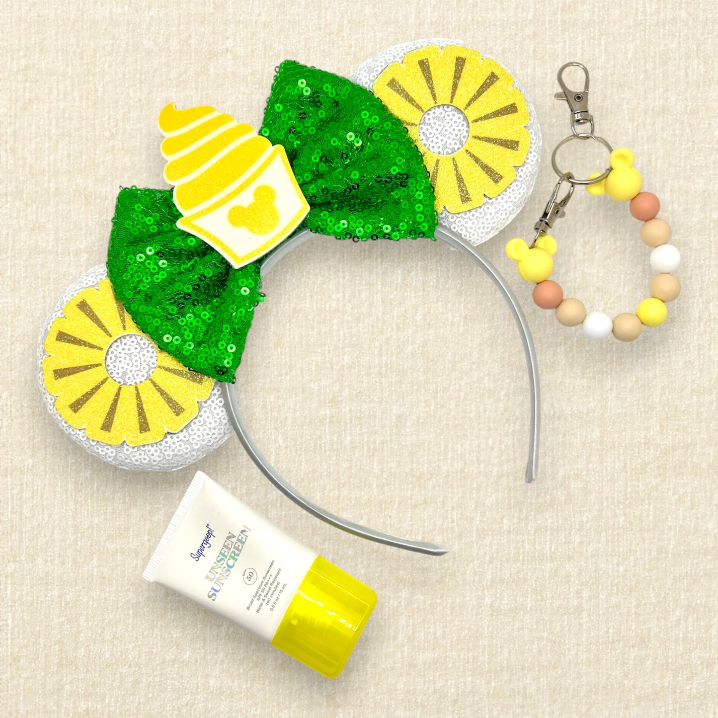 White mouse ears with glitter pineapple rings, green bow, and cup of pineapple swirl soft serve; sunscreen; yellow beaded mouse ears holder clip