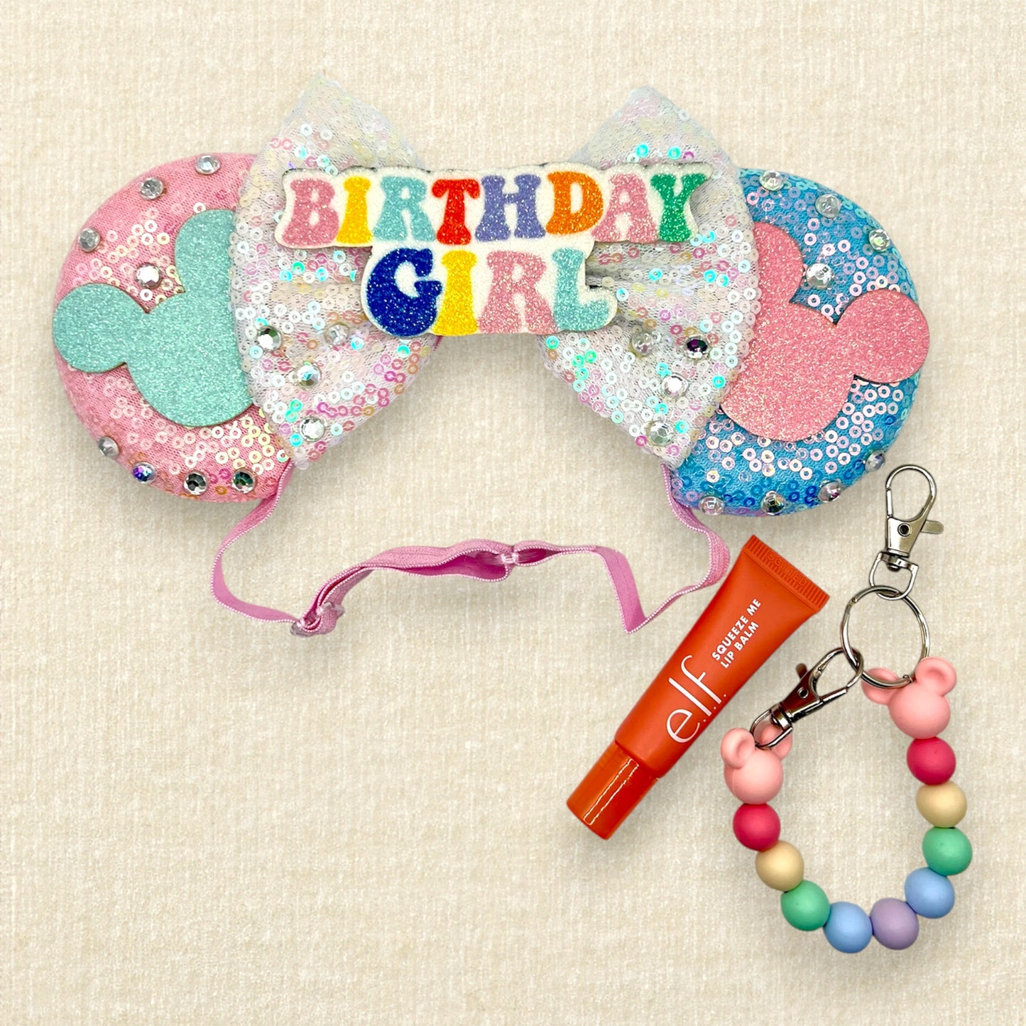Pink and blue mouse ears with glitter mouse silhouettes, iridescent white sequin bow, and "Birthday Girl" text centerpiece; pink lip gloss; beaded mouse ears holder