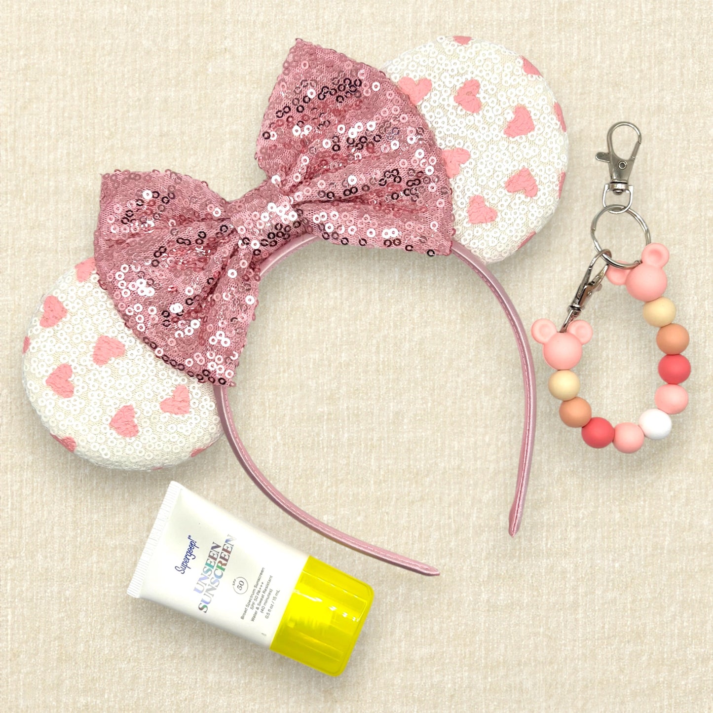 White sequin mouse ears with small pink hearts and rose gold sequin bow; small tube of sunscreen; pink beaded mouse ears holder