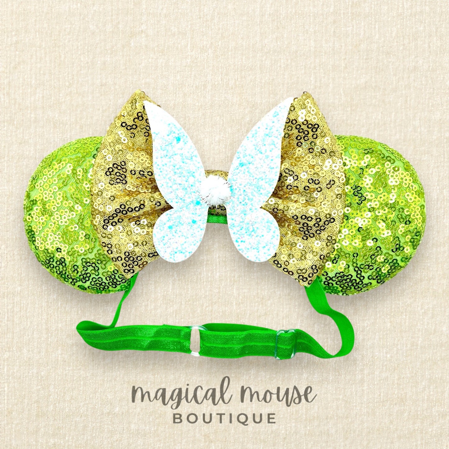 Green sequin mouse ears headband with gold bow and glitter fairy wings, on a beige background with "Magical Mouse Boutique" branding
