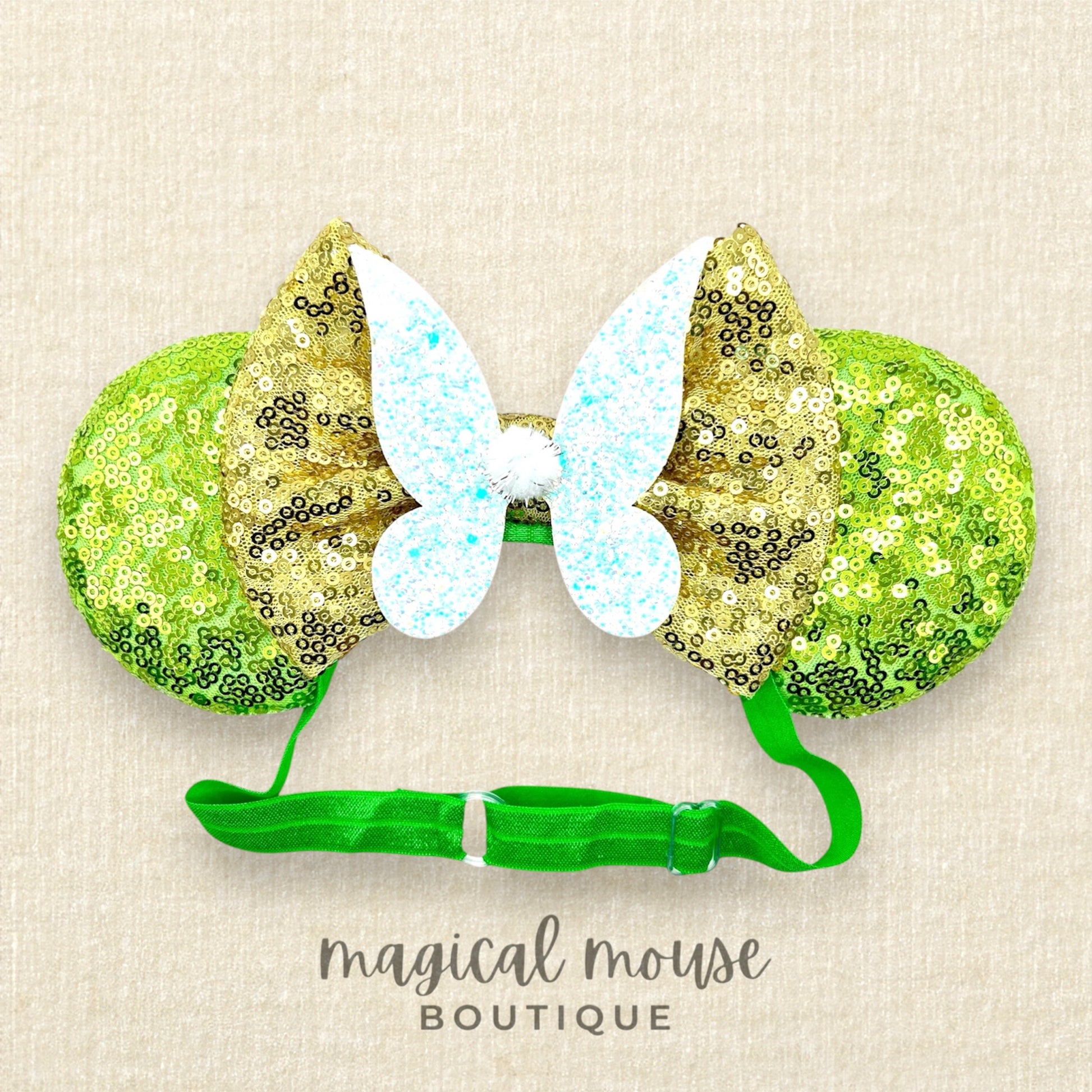 Green sequin mouse ears headband with gold bow and glitter fairy wings, on a beige background with "Magical Mouse Boutique" branding