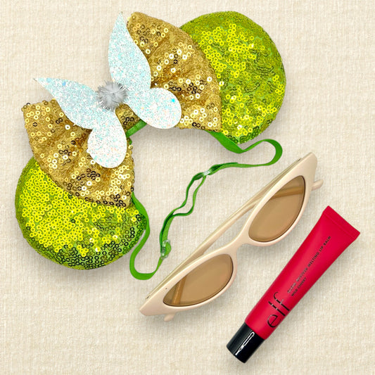 Light green sequin mouse ears with gold sequin bow and glitter fairy wings on an elastic headband; cream cat-eye sunglasses; tube of red lip gloss