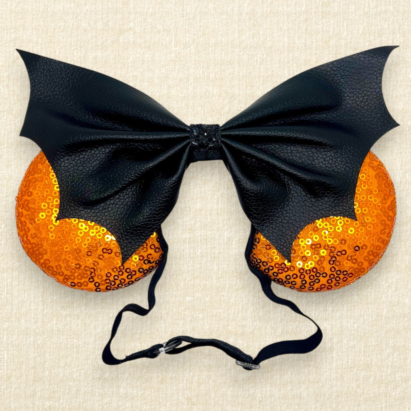 Halloween-themed mouse ears with orange sequin ears and black faux leather bat wing bow on an elastic headband