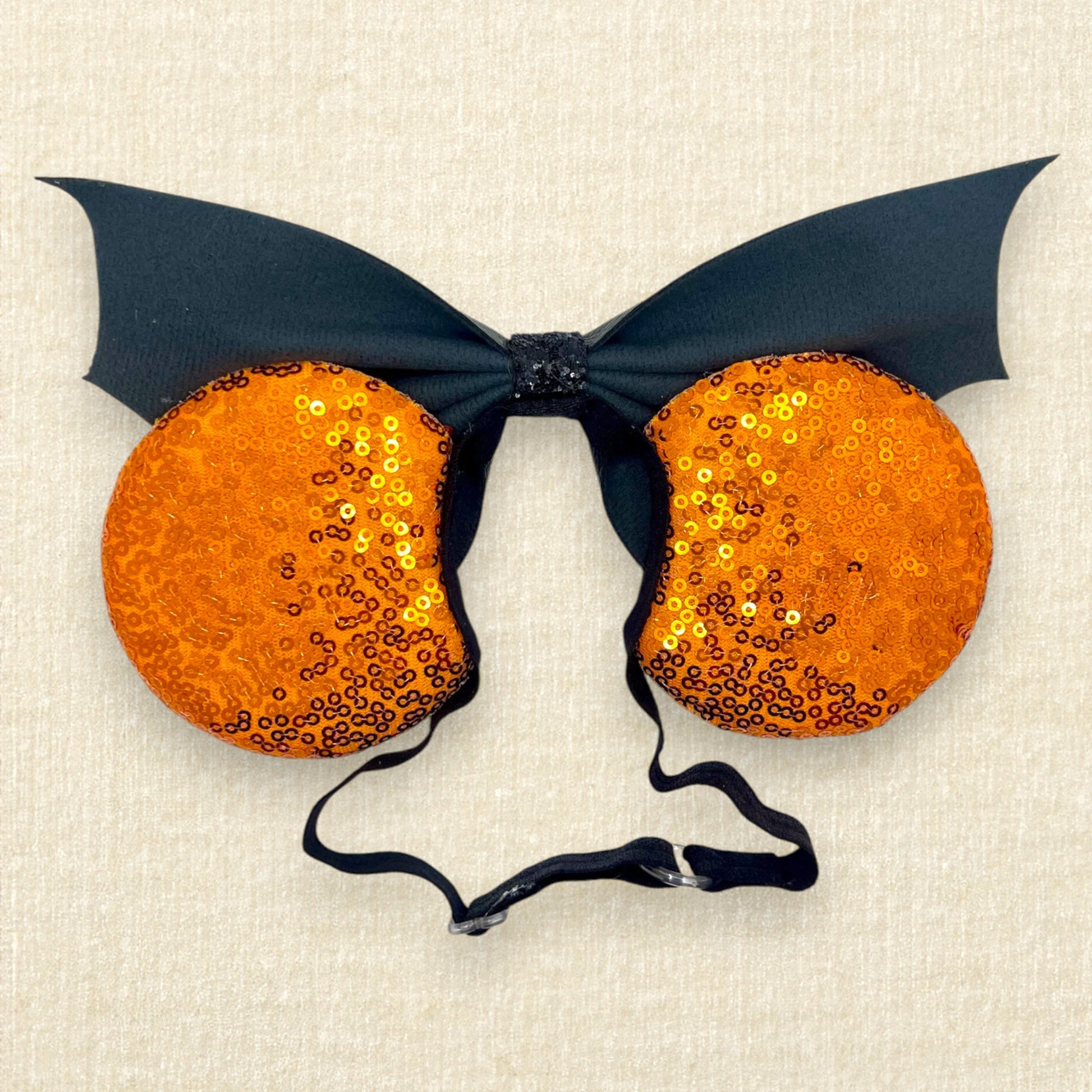 Halloween-themed mouse ears with orange sequin ears and black faux leather bat wing bow on an elastic headband (back view)