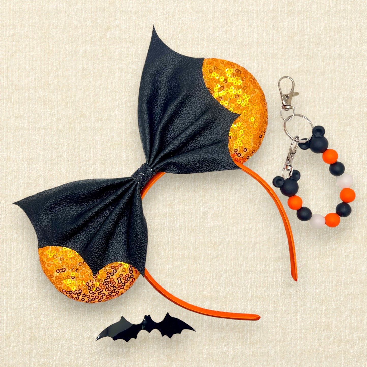 Halloween-themed mouse ears headband with orange sequin ears and black faux leather bat wing bow; black bat decoration; black and orange beaded mouse ears holder keychain