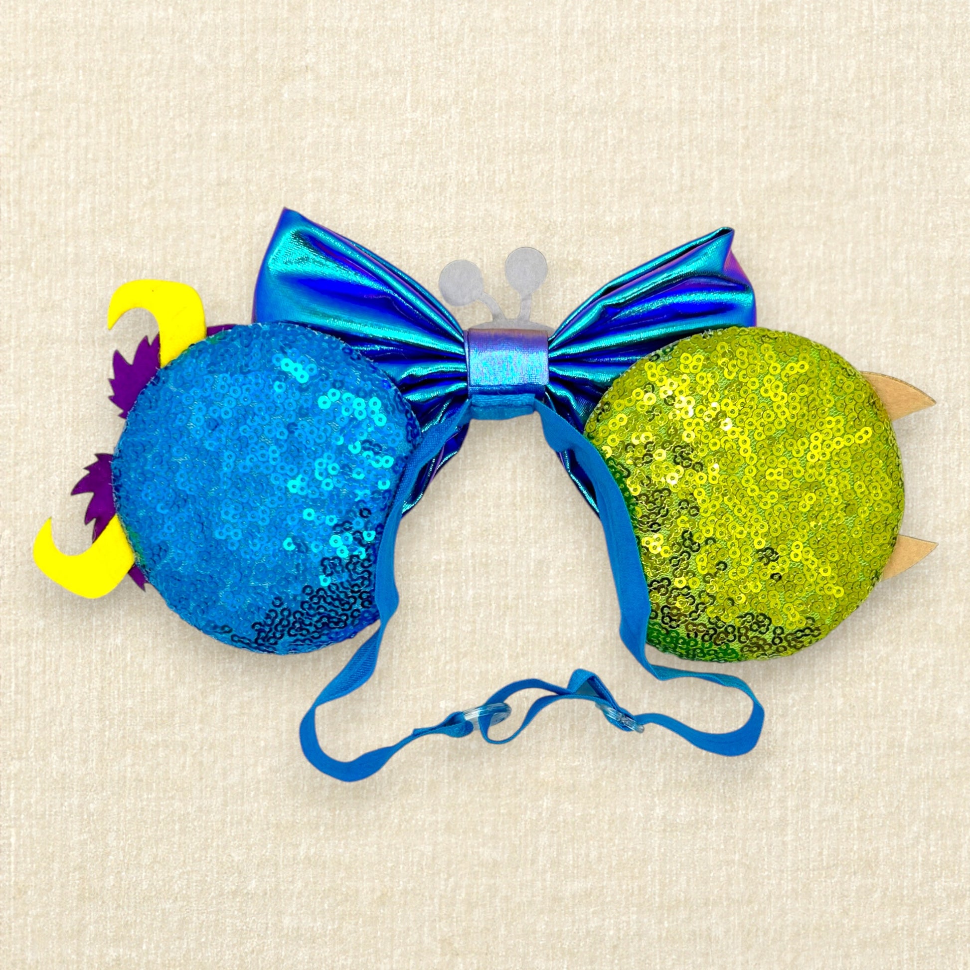 Green and blue sequin mouse ears with glitter monster details and an iridescent blue bow on an elastic headband (back view)